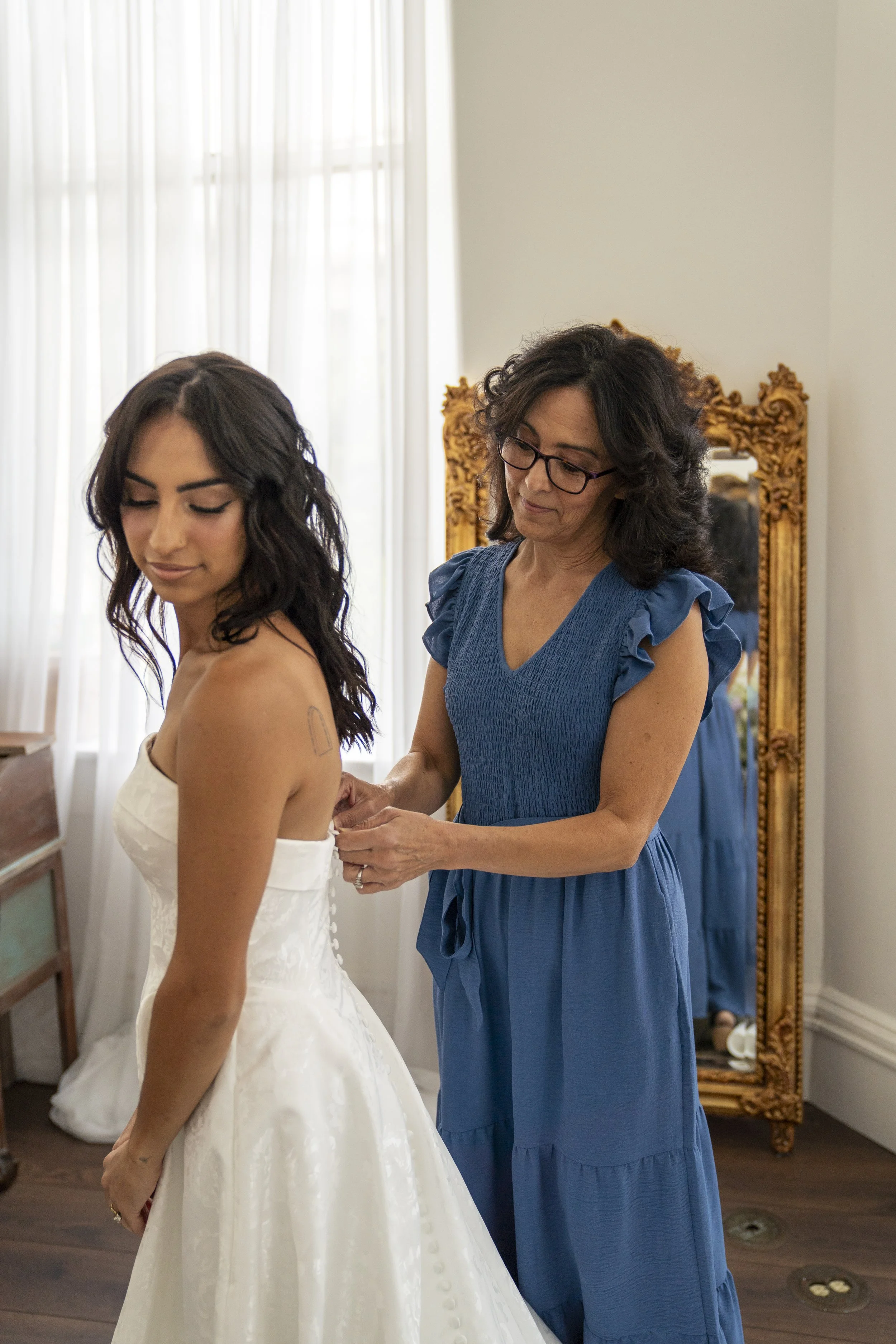 A woman with long dark hair in a white wedding dress stands with her eyes closed while a woman in a blue dress helps her, possibly getting ready for a wedding, in a room with sheer curtains and a large ornate mirror.