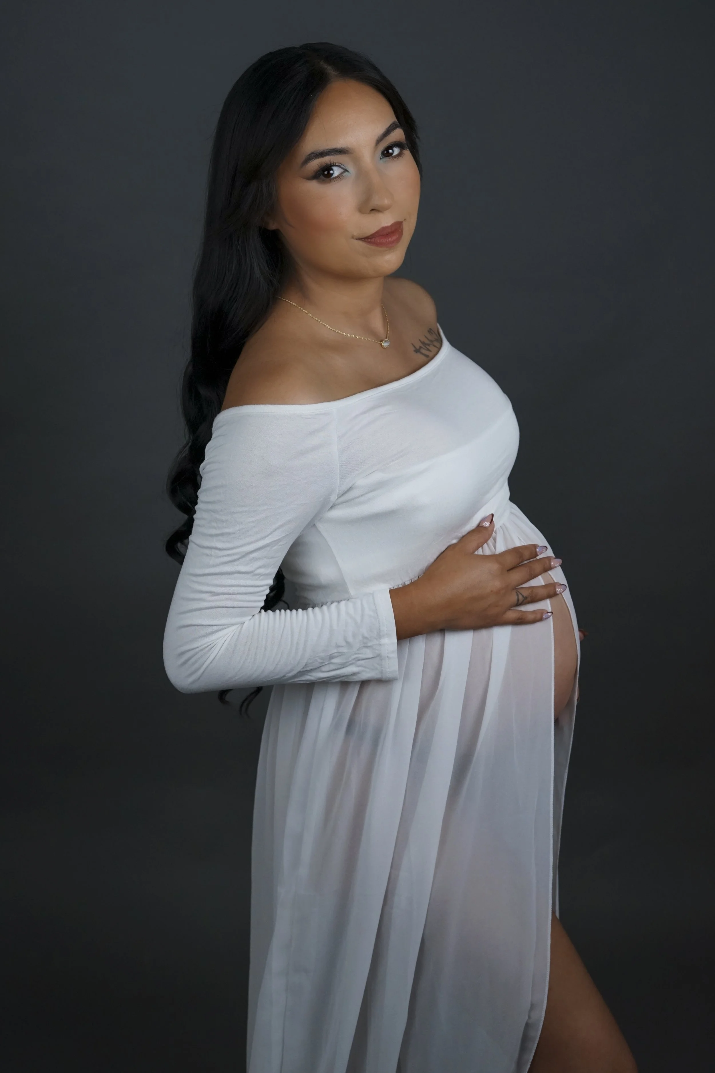 A pregnant woman with long dark hair, wearing a white off-shoulder dress, standing against a dark background.