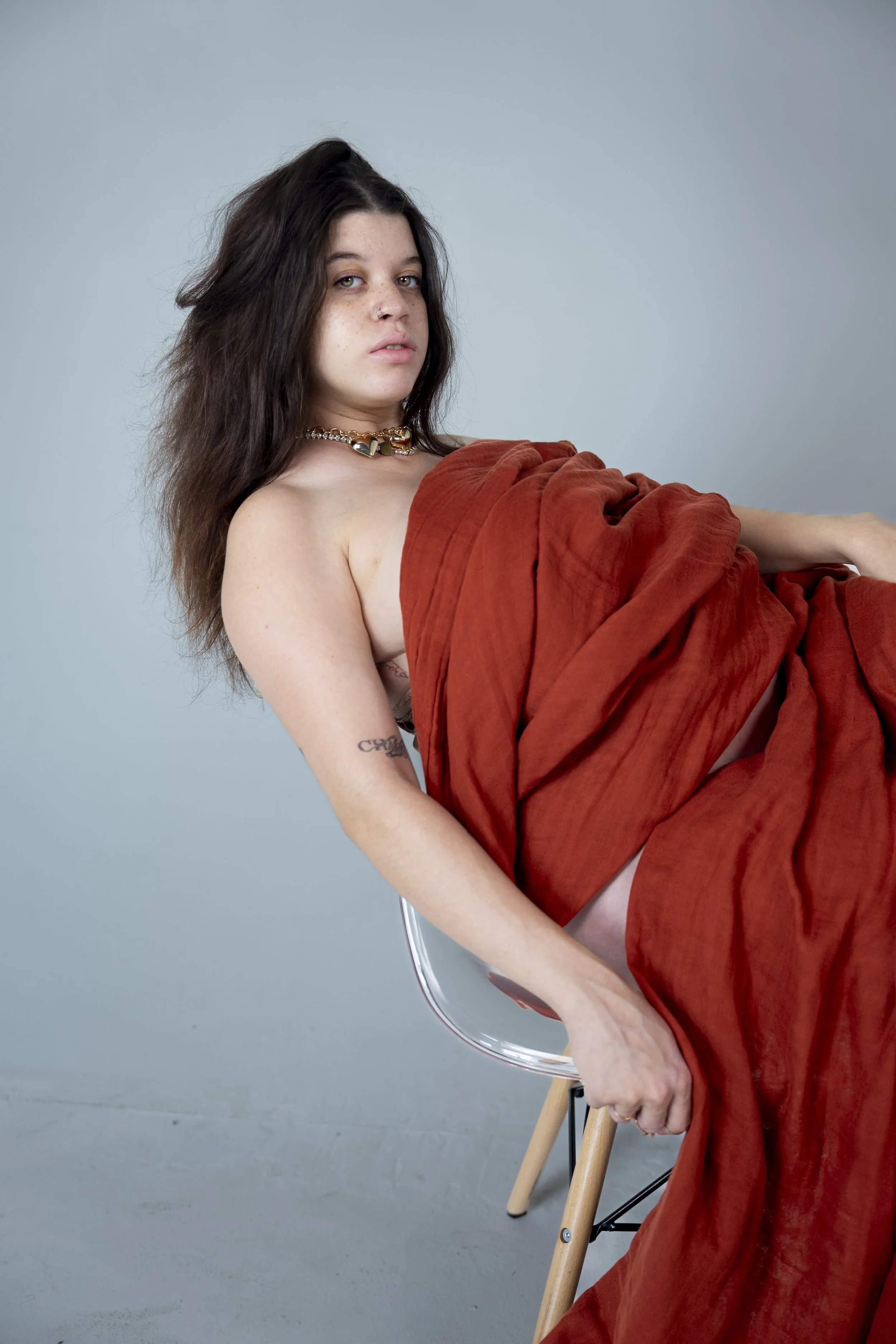 Young woman with dark, slightly curly hair, sitting on a transparent chair with wooden legs, draped with a rust-colored fabric, against a plain light blue background.