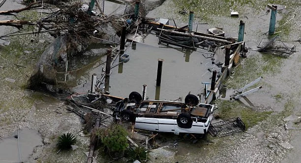 An overturned white vehicle submerged in floodwaters with debris and damaged structures around, indicating severe flooding and destruction.