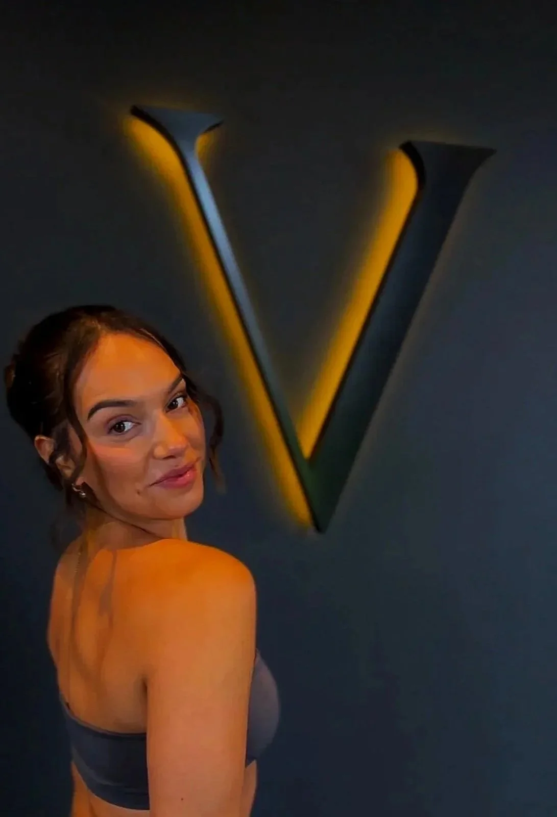 A woman standing in front of a large illuminated letter V on a dark wall.