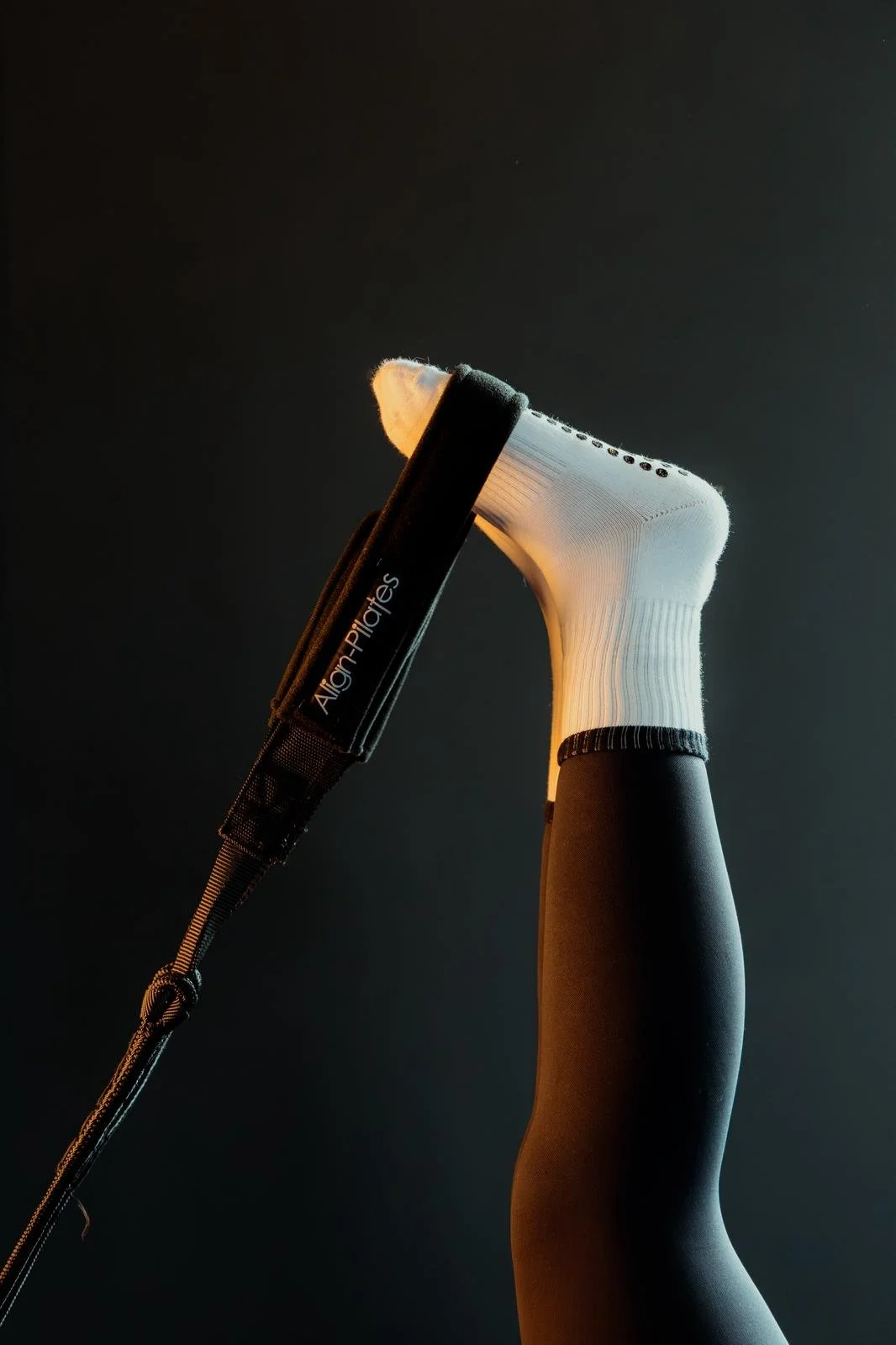 A closeup of someone's leg wearing black leggings and a white grip sock. The foot is in a reformer pilates leg strap and the leg is in the air.