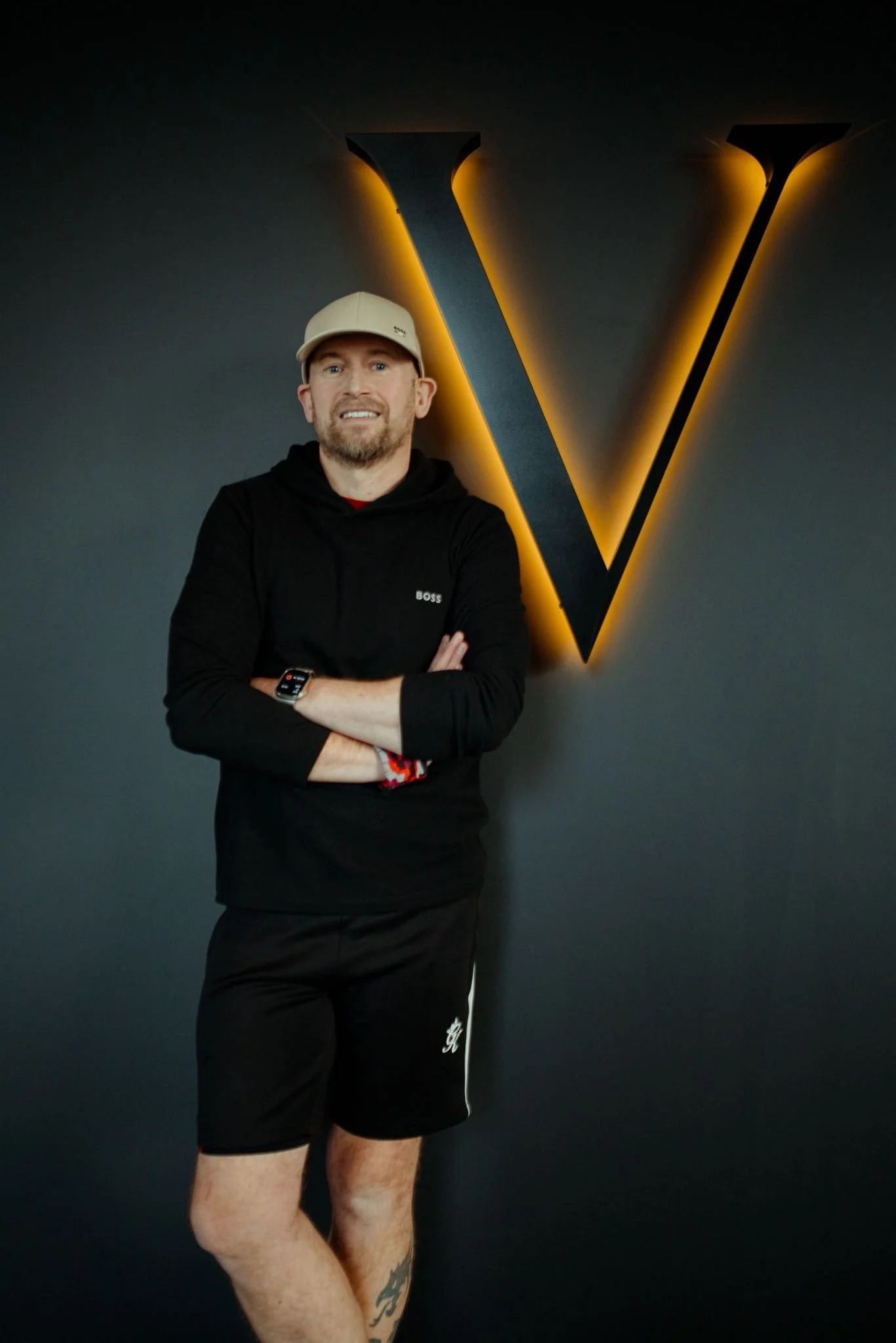 A white male reformer pilates instructor is standing with his arms crossed and smiling at the camera. He's wearing all black and a cream baseball cap. He is standing in front of a brand wall at Vitalis Studios. There is a big V on the wall.