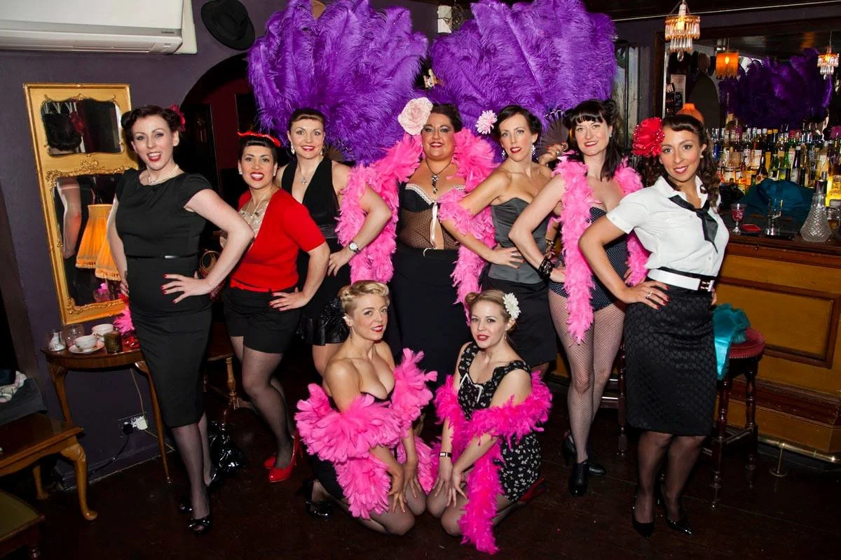 A group of nine women in vintage pin-up style costumes posing together in a bar or restaurant. Some women are wearing feather boas and 1940s inspired clothing, with bright smiles and glamorous makeup. The background has bar shelves with bottles and purple feather decorations.