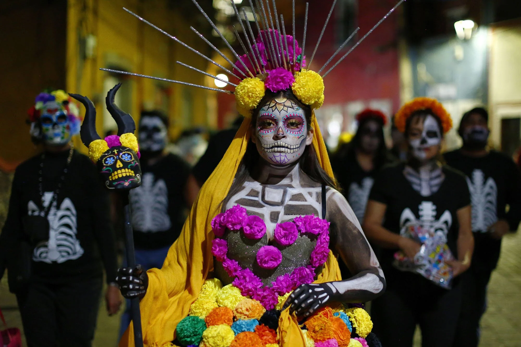 A woman dressed in a Day of the Dead costume with colorful face paint resembling a skull, wearing a yellow dress decorated with pink and orange flowers, and a large headpiece with pink, yellow, and purple flowers and silver spikes. She is holding a bouquet of colorful flowers, with other similarly dressed individuals in the background.