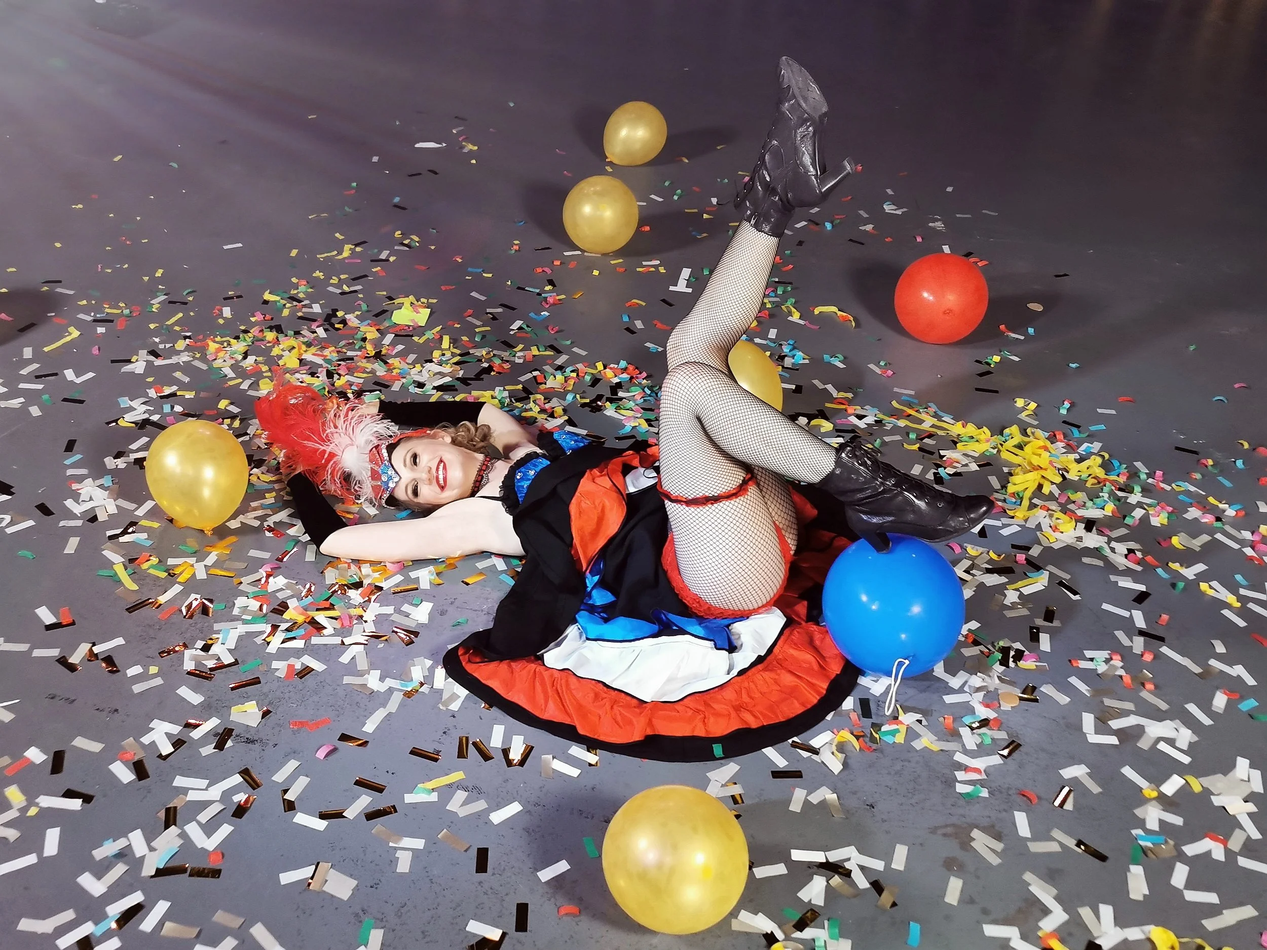A woman dressed in a clown costume lying on the floor surrounded by colorful confetti and balloons.