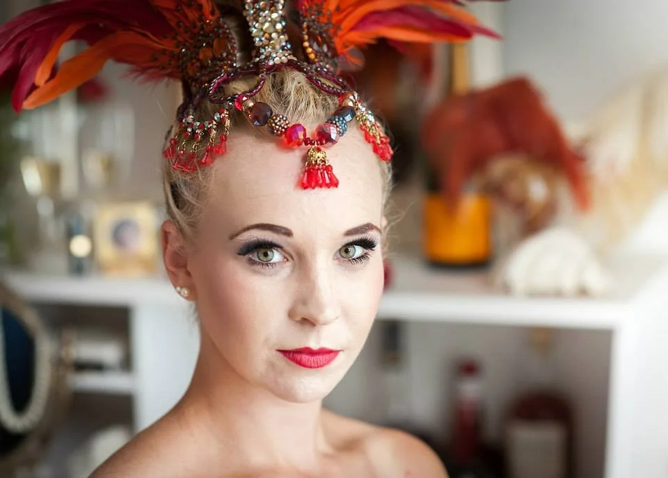 A woman with light skin, blonde hair, and blue eyes wearing elaborate headwear with red feathers and beaded accessories. She has makeup with red lipstick and eye makeup, and is looking directly at the camera.