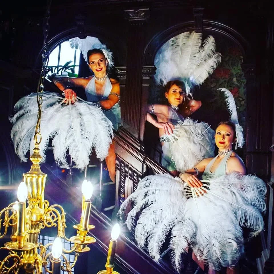 Three women dressed in glamorous outfits with feather fans, posing on a staircase in a fancy setting.