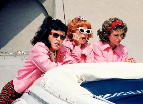 Three women in pink shirts with sunglasses and headbands leaning on a white vehicle, with a building in the background.
