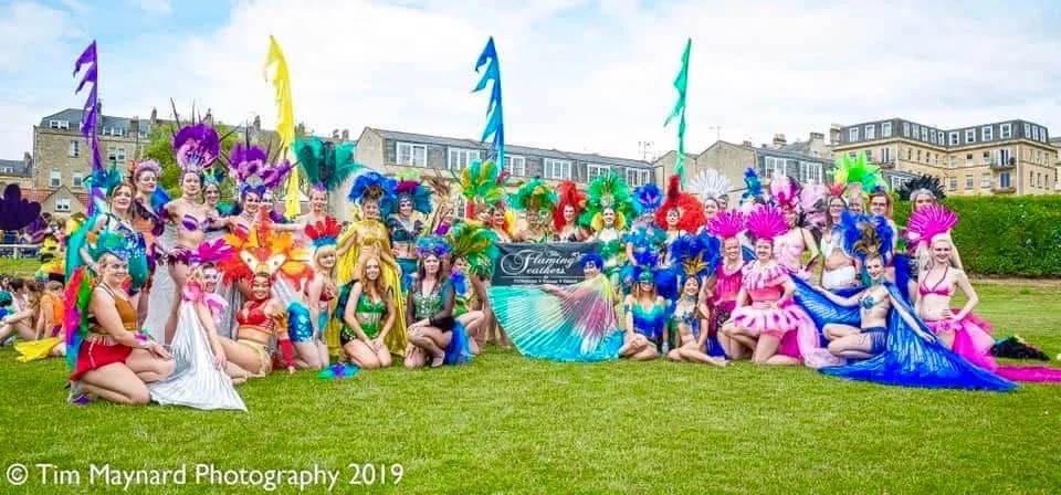 Group of women in colorful, elaborate carnival costumes with feathers, sequins, and boas, posing outdoors on a grassy field during daytime.