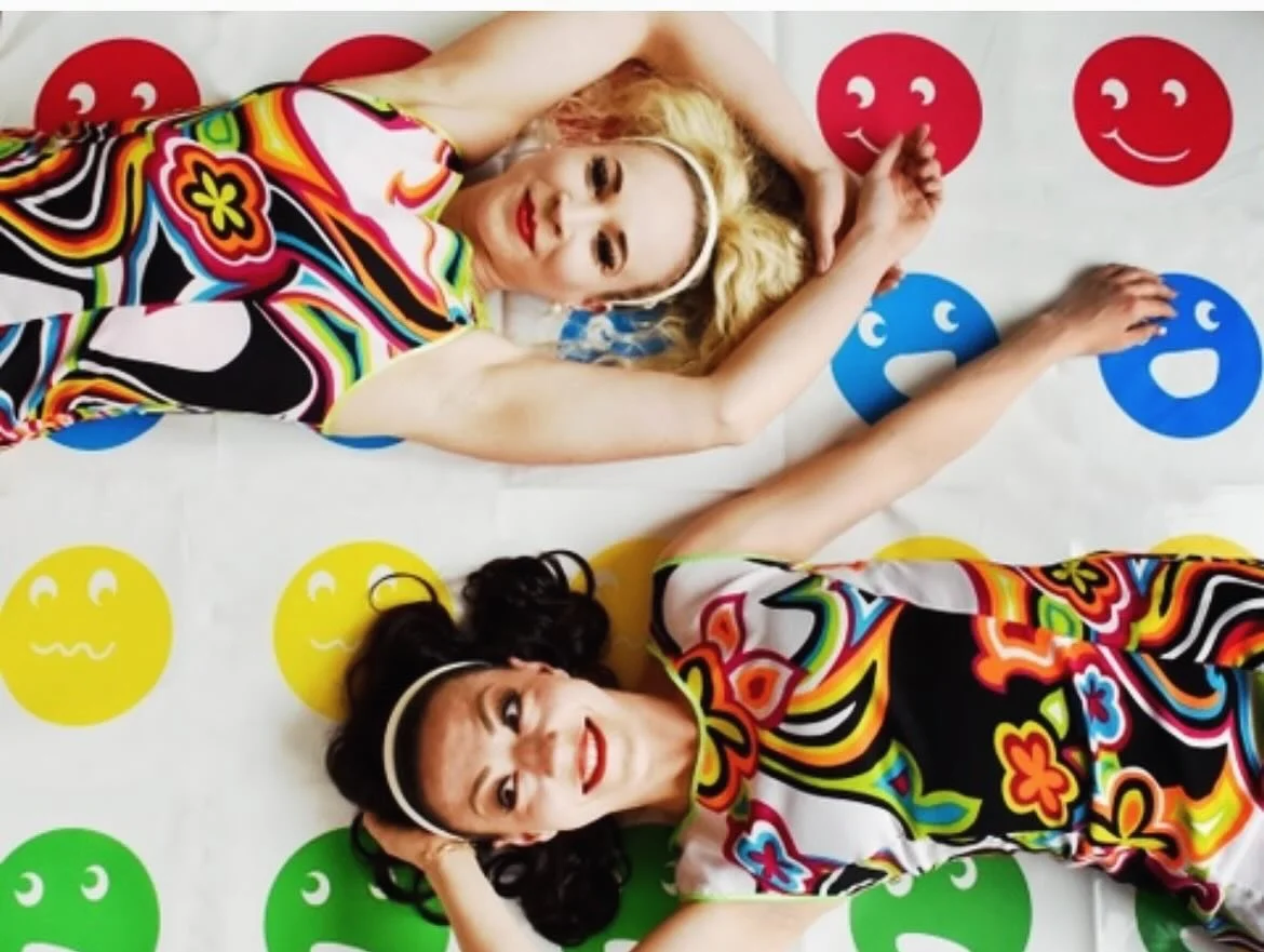 Two women lying on a colorful smiley face floor mat, wearing vibrant, patterned dresses and smiling. One has blonde hair with a white headband, the other has dark hair with a dark headband. They are looking up at the camera with cheerful expressions.