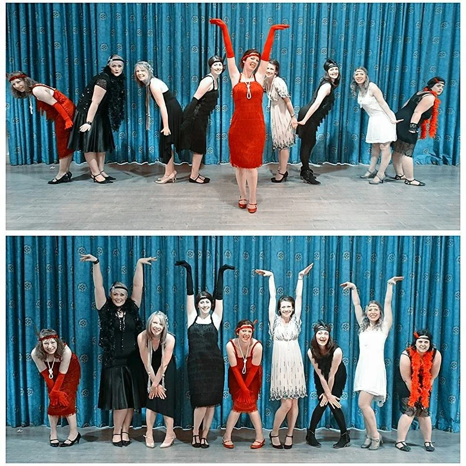 Group of women dressed in 1920s-style fashion, posing on a stage with a blue curtain backdrop, in vintage flapper dresses, headbands, and accessories, likely performing or celebrating a themed event.