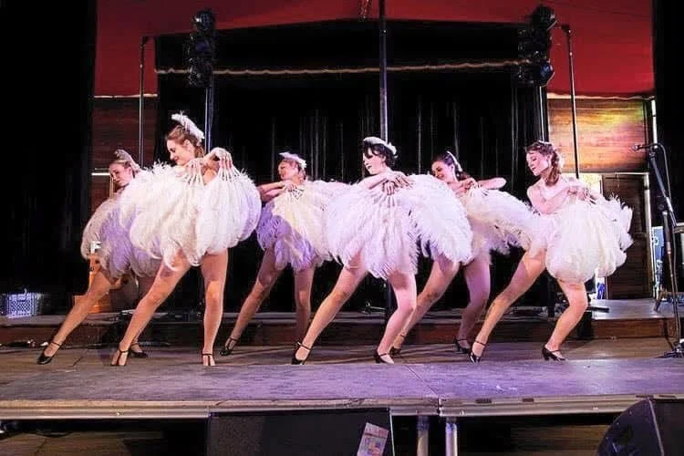 Group of six women performing a dance on stage in white, feathered costumes with black high heels, in front of a black curtain.