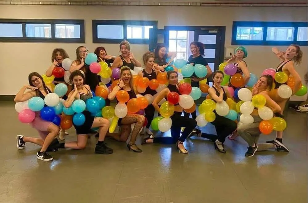 A group of women wearing dance costumes with colorful balloons attached, posing playfully in a studio or gym. They are smiling and striking various poses.