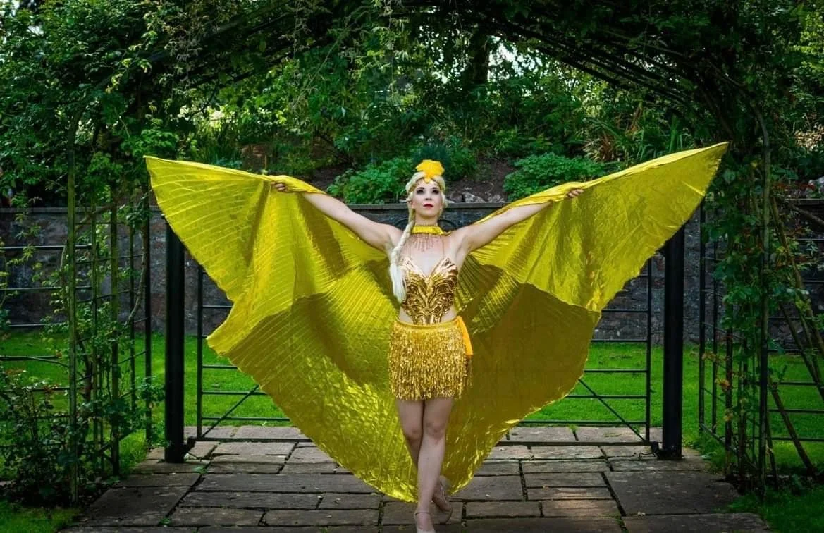 A woman standing outdoors with her arms extended, wearing a golden dress and large yellow butterfly wings.
