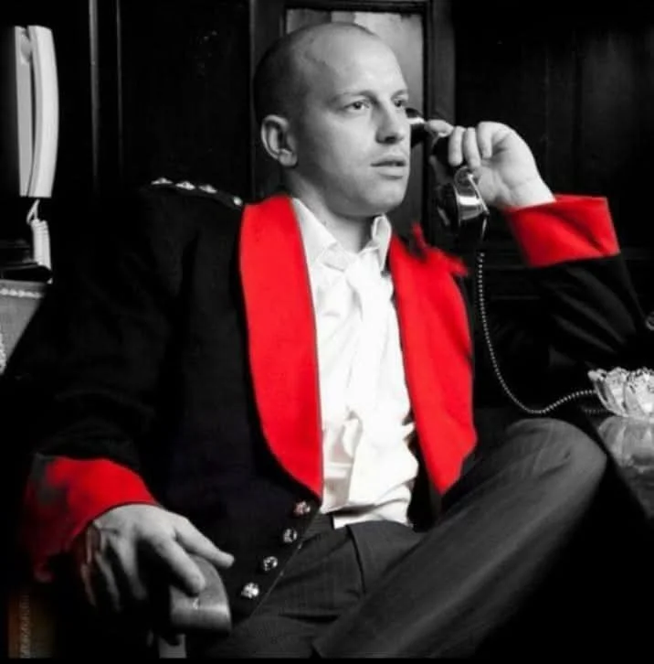 A man in a black jacket with red cuffs and collar, white shirt, sitting on a chair, holding a phone to his ear, with a serious expression.