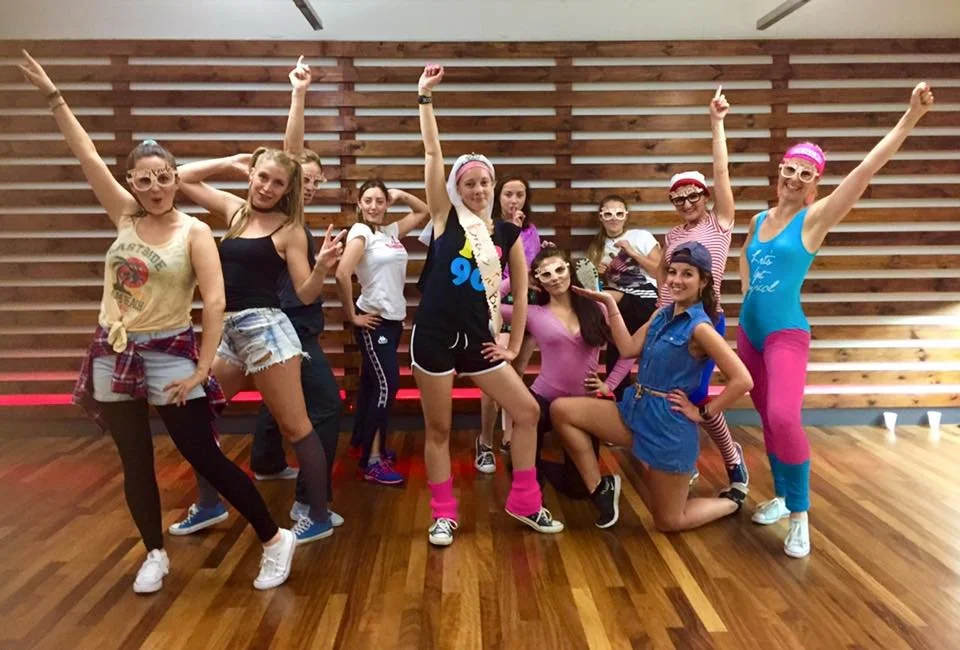 Group of ten women dressed in colorful workout or dance costumes, posing with energetic and fun gestures in a room with wooden flooring and a wooden slat wall.