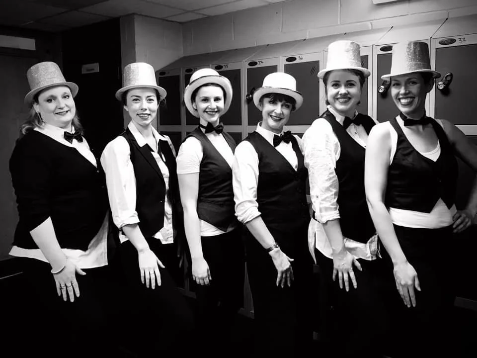 Seven women dressed as waitresses or performers in uniform with bow ties, vests, and hats, standing in a row in front of lockers in a backstage area, smiling at the camera.