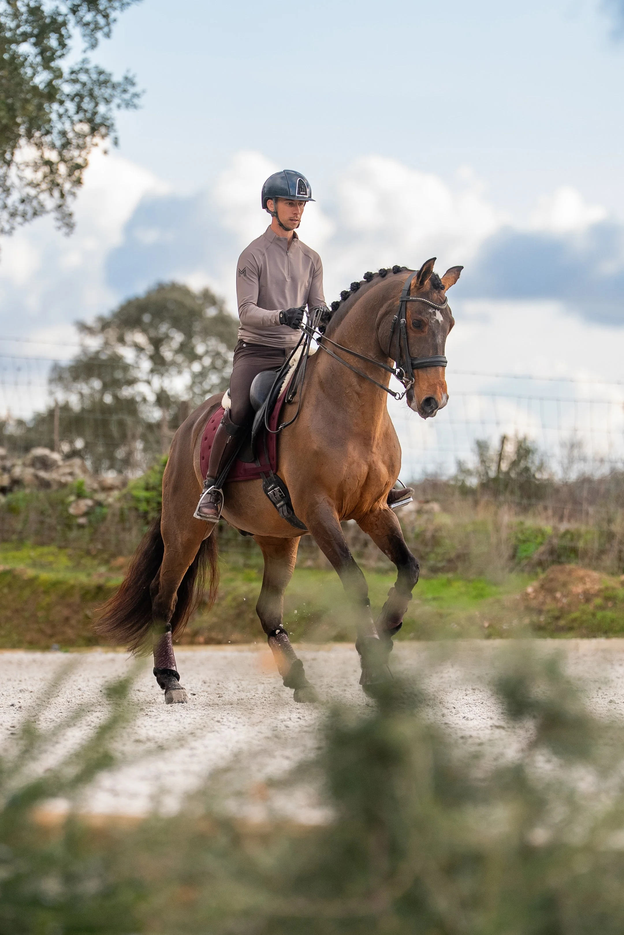 A male equestrian rider in a gray jacket and black helmet riding a brown horse on a gravel path outdoors with trees and cloudy sky in the background.