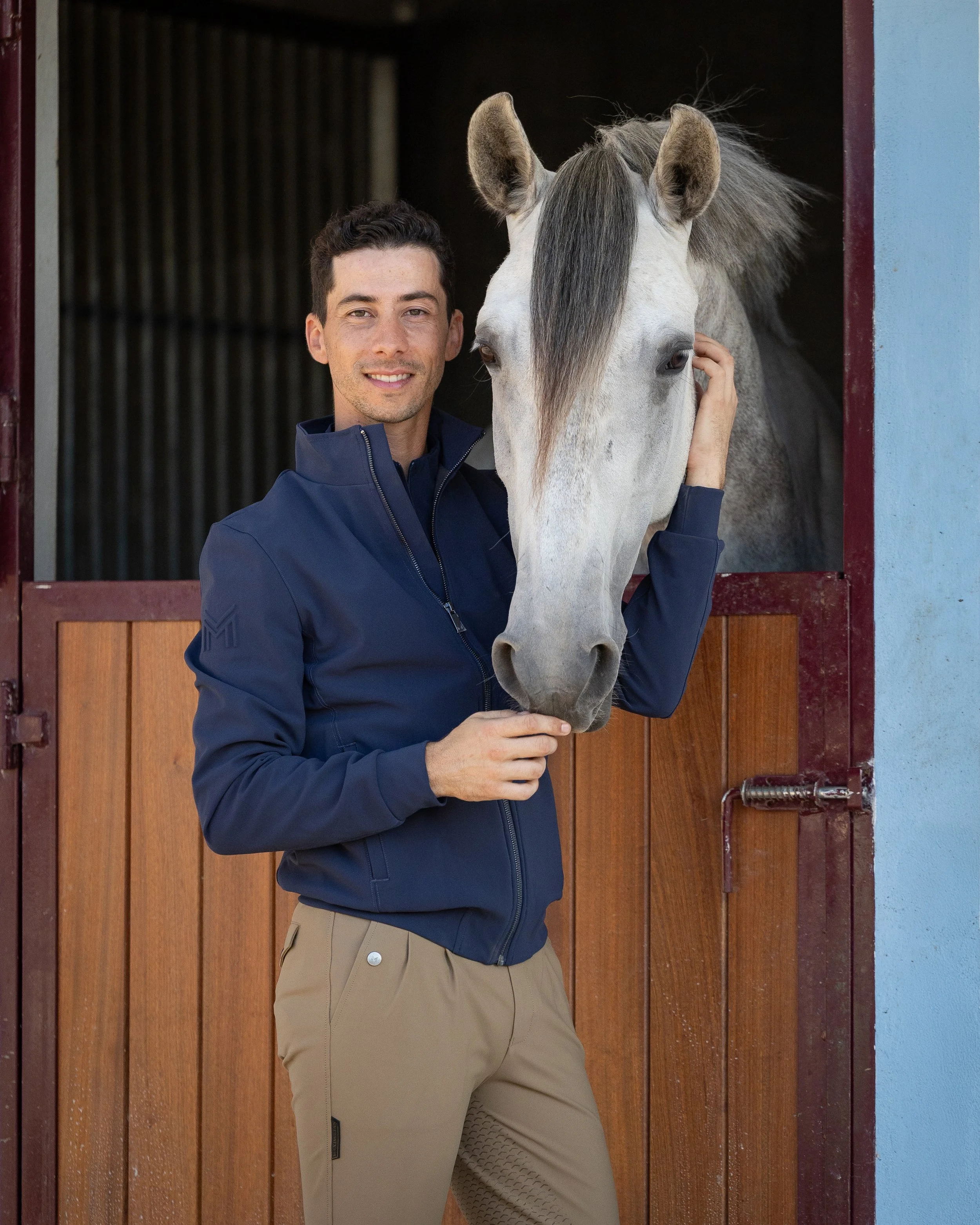 A young man standing next to a gray horse, smiling, with his hand on the horse's face, in front of a wooden stable door.