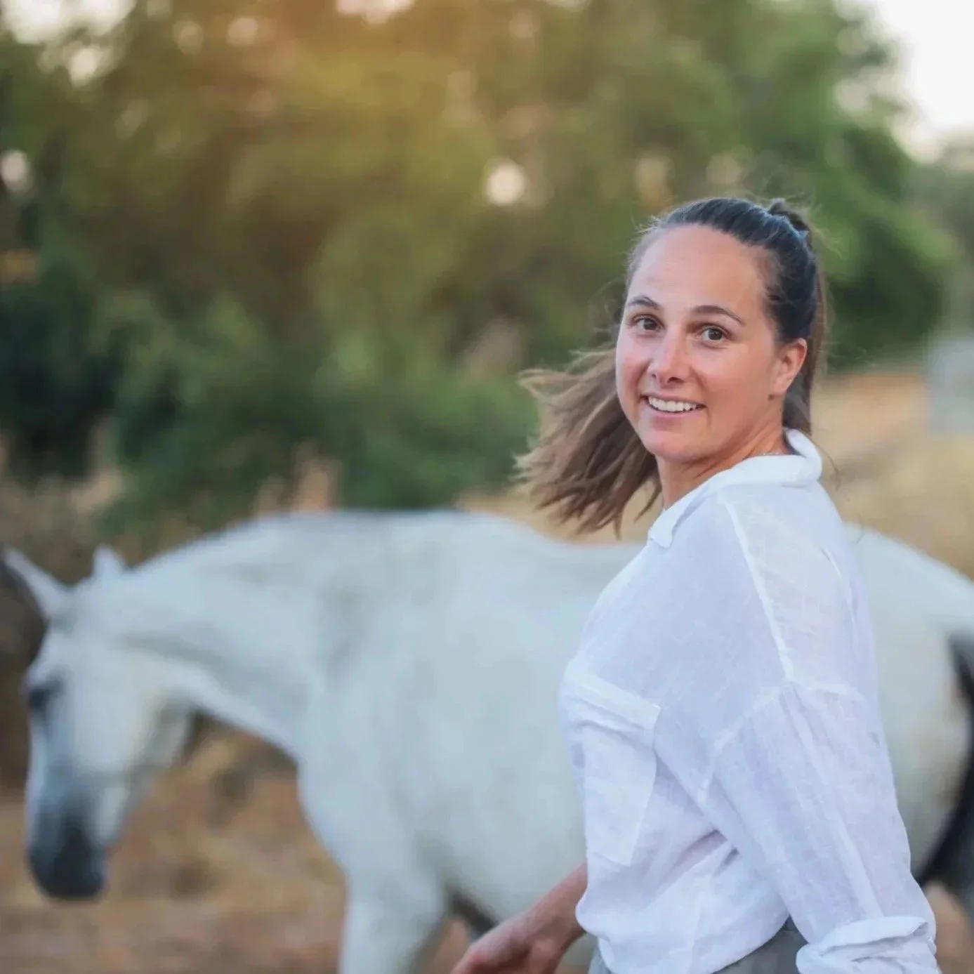 Woman with brown hair in a ponytail wearing a white shirt, smiling, standing outdoors with a white horse in the background.