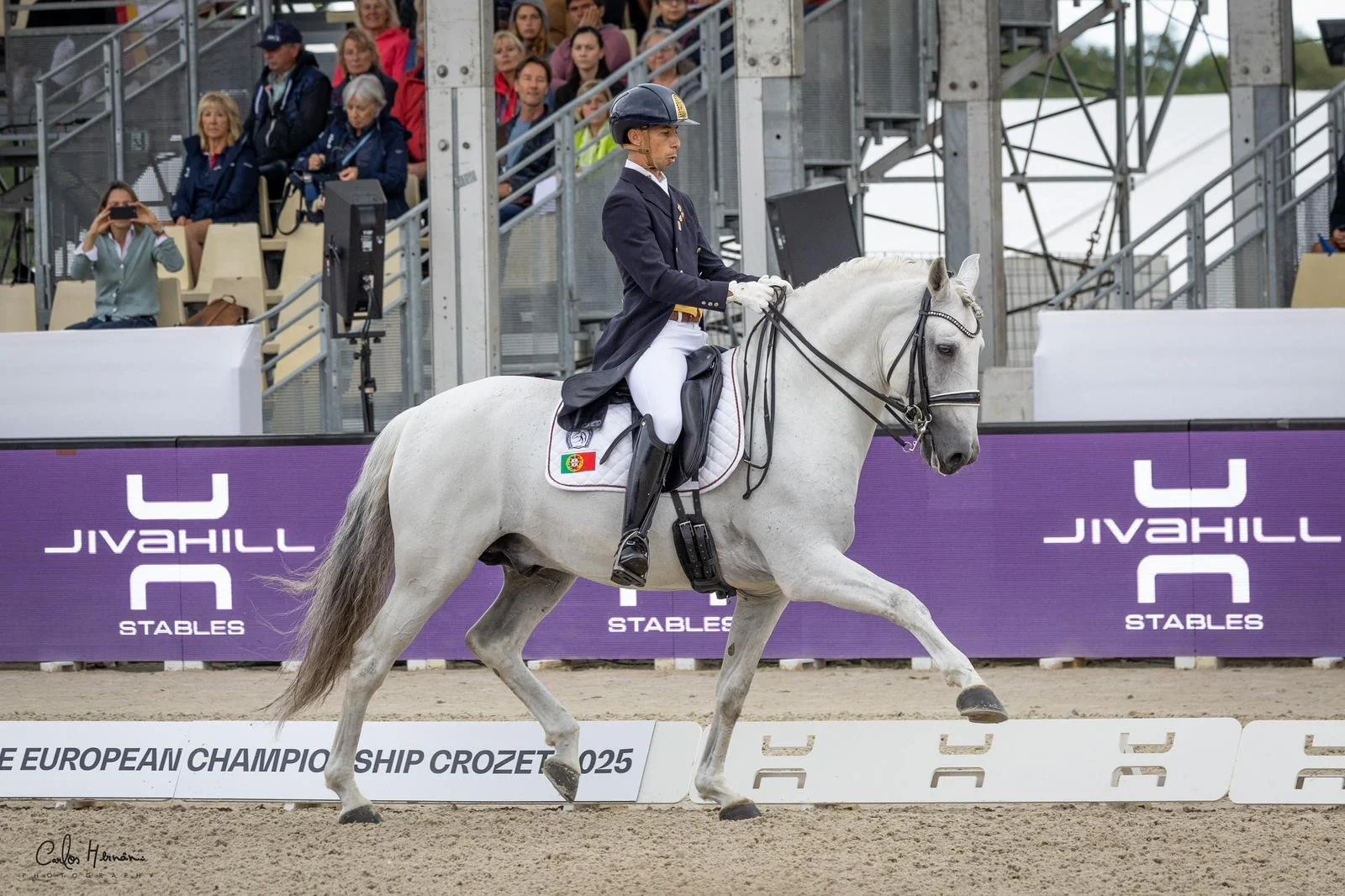 A male equestrian rider in formal riding attire riding a white horse during a dressage competition at the European Championship Crozet 2025, with spectators in the background.
