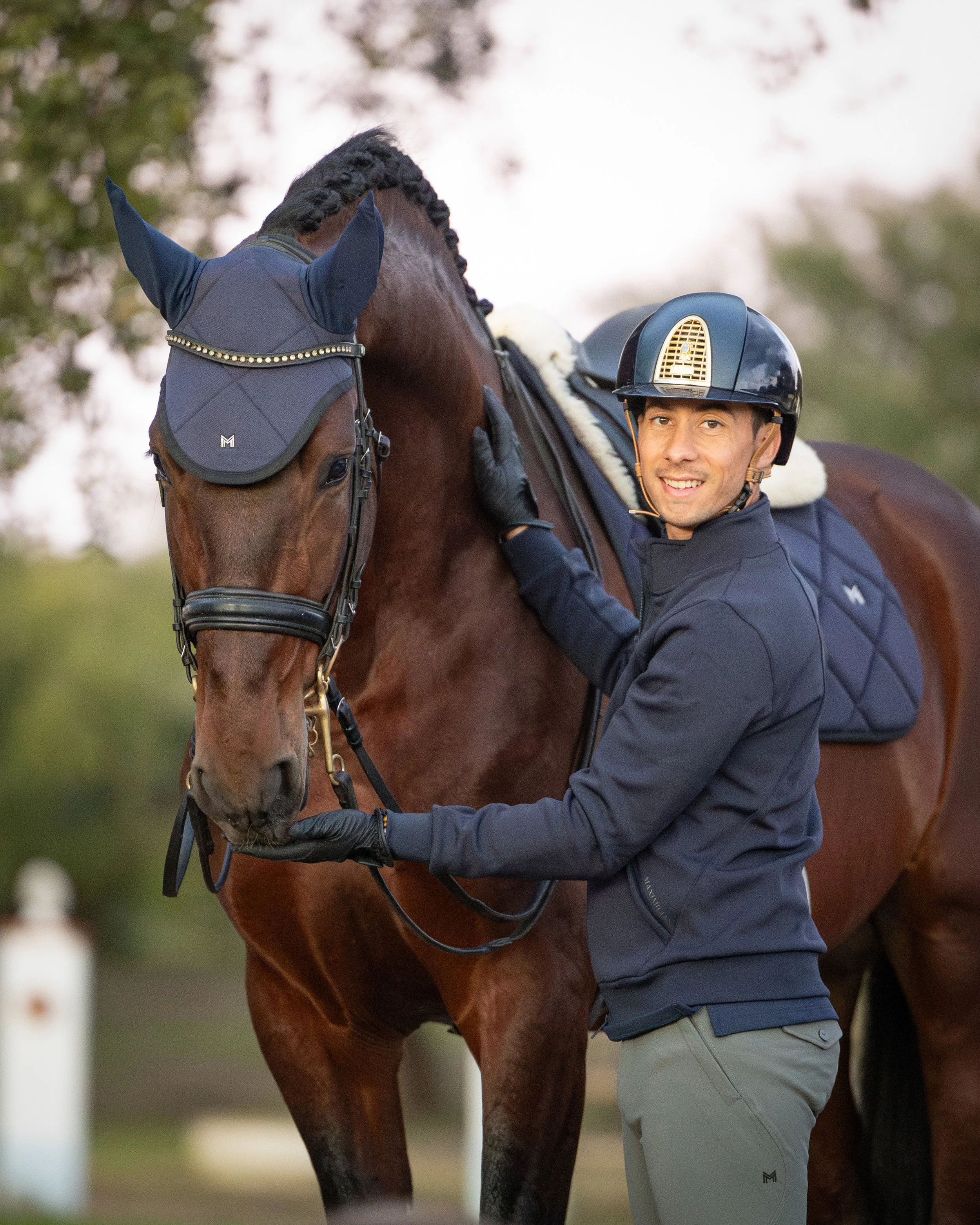 A man in riding gear smiles and holds a horse's reins, standing beside his horse outdoors.