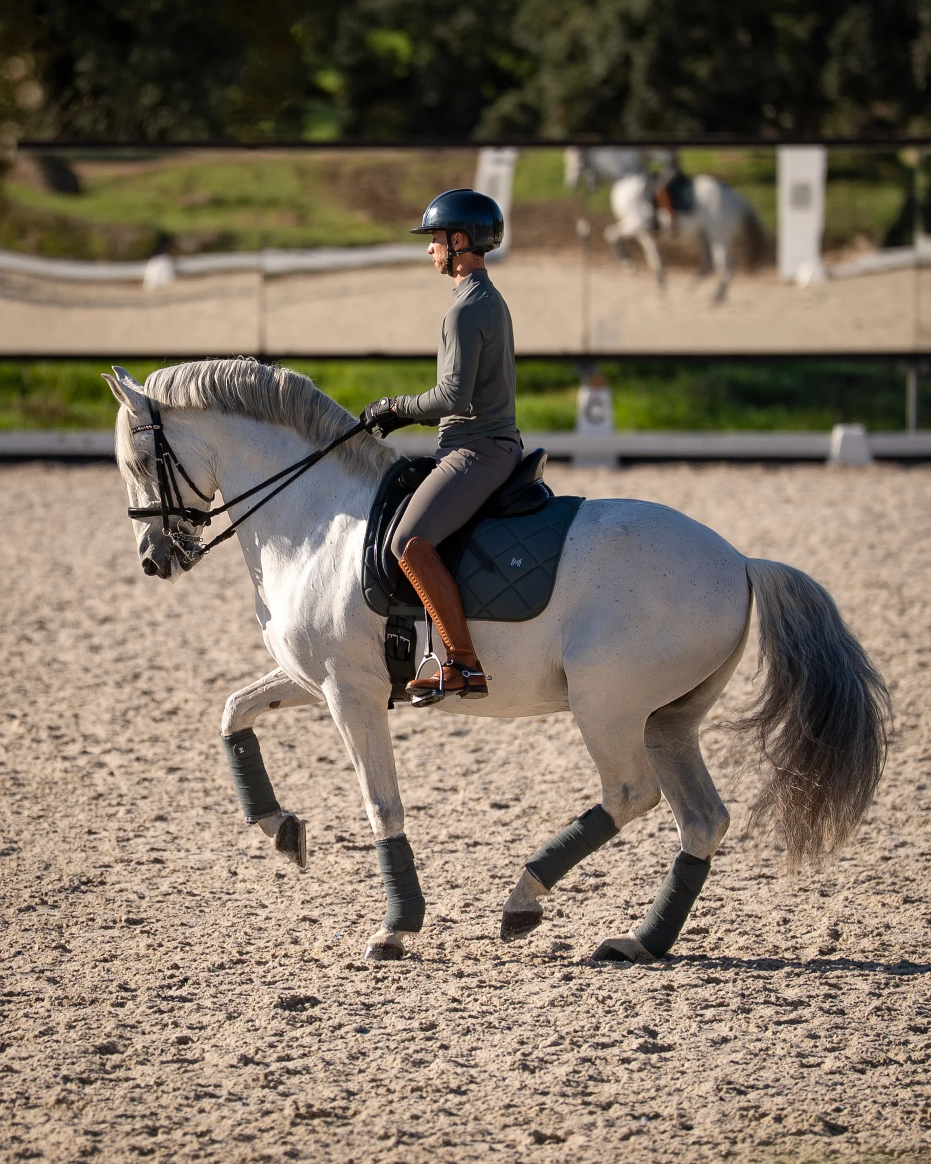 A woman riding a white horse on an outdoor riding arena, with a mirror wall reflecting the rider and horse.