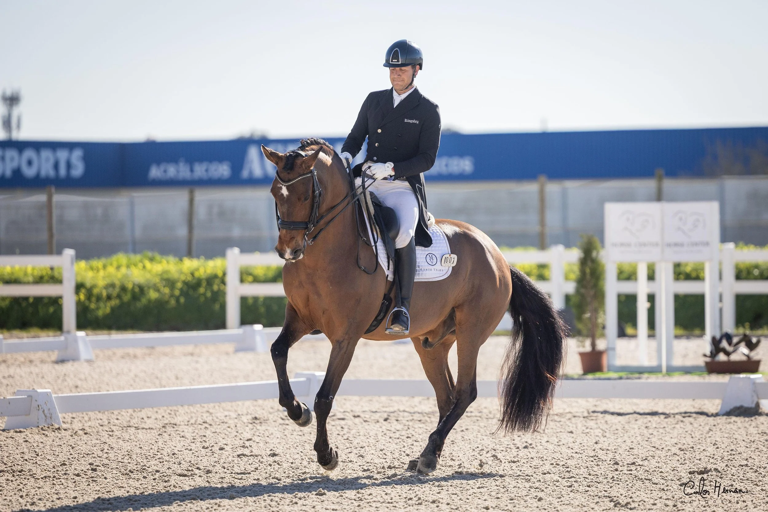 A man riding a horse in a dressage arena during a competition under a clear sky.