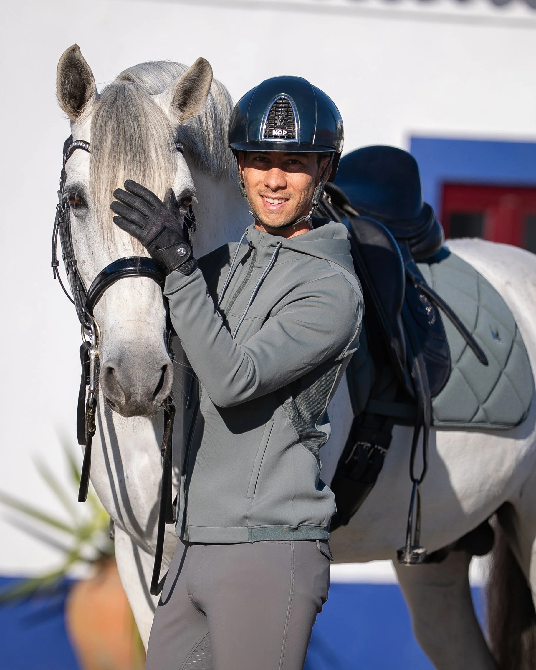 A man in riding gear and helmet stands beside a white horse with a gray mane, touching its face while smiling at the camera.
