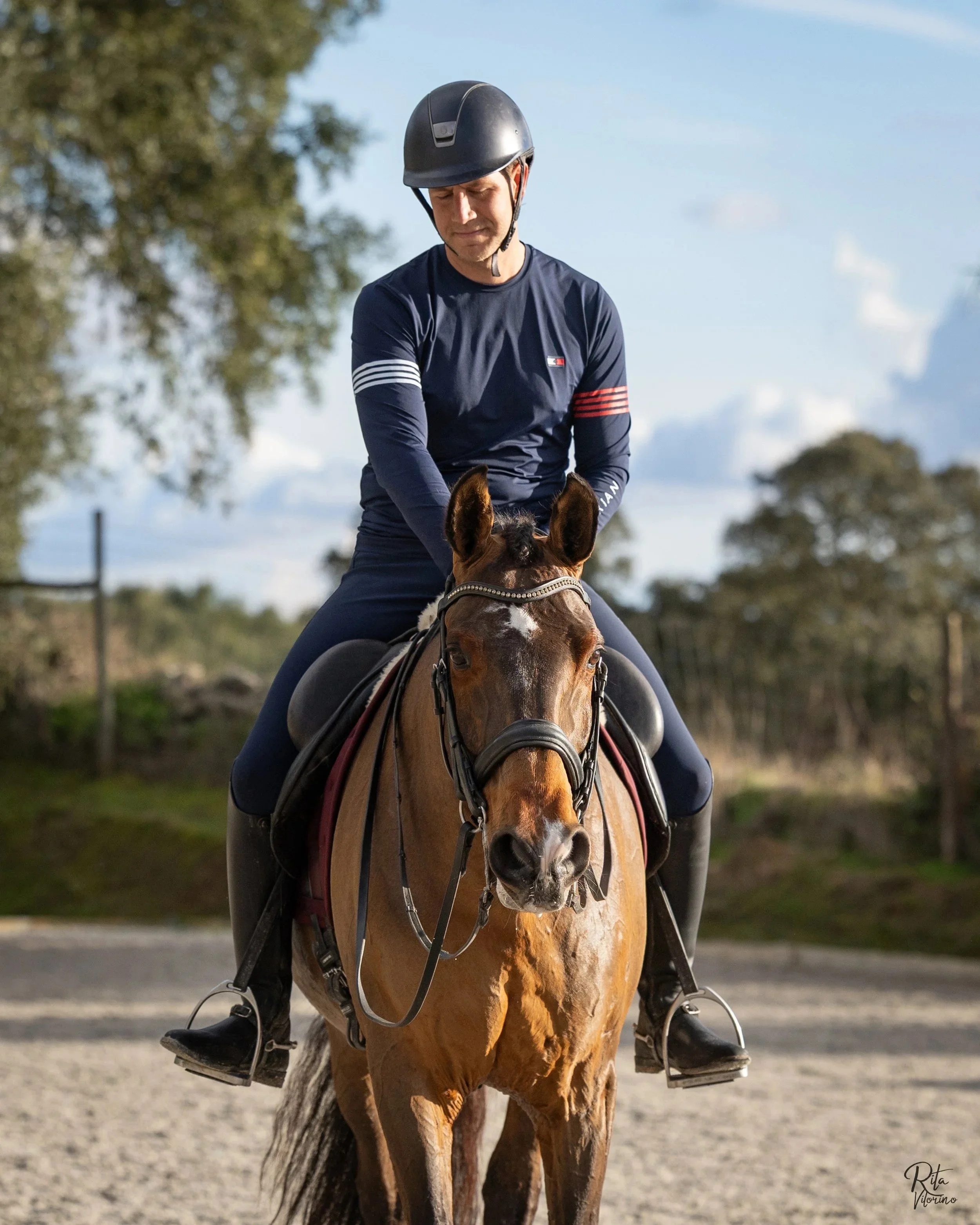 A man wearing a black riding helmet and navy long-sleeve shirt riding a brown horse outdoors with trees and blue sky in the background.