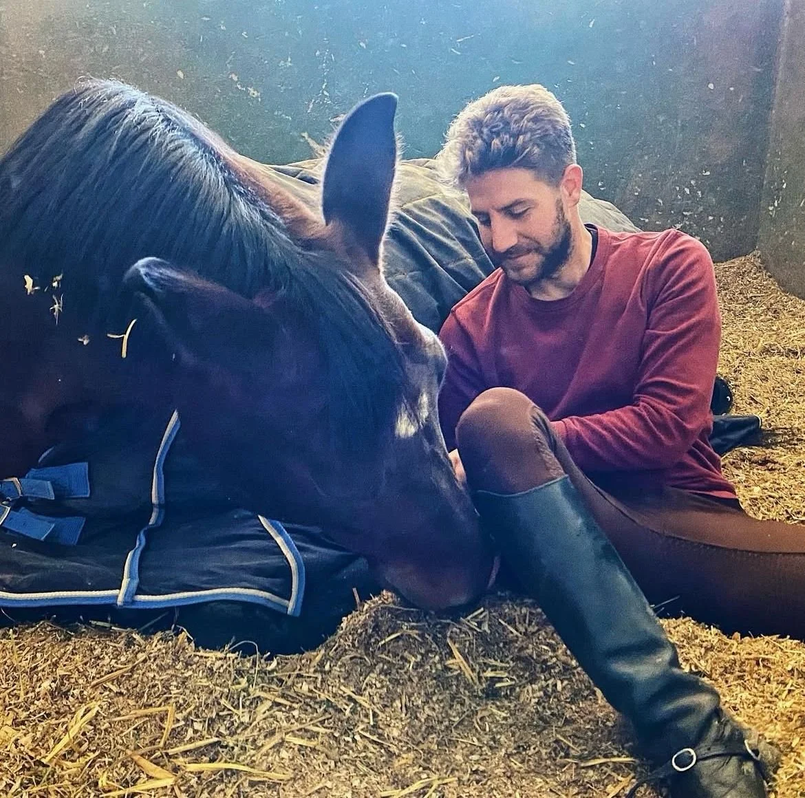 A man sitting on hay with a horse, leaning towards each other with casual, relaxed expressions, in a stable-like environment.