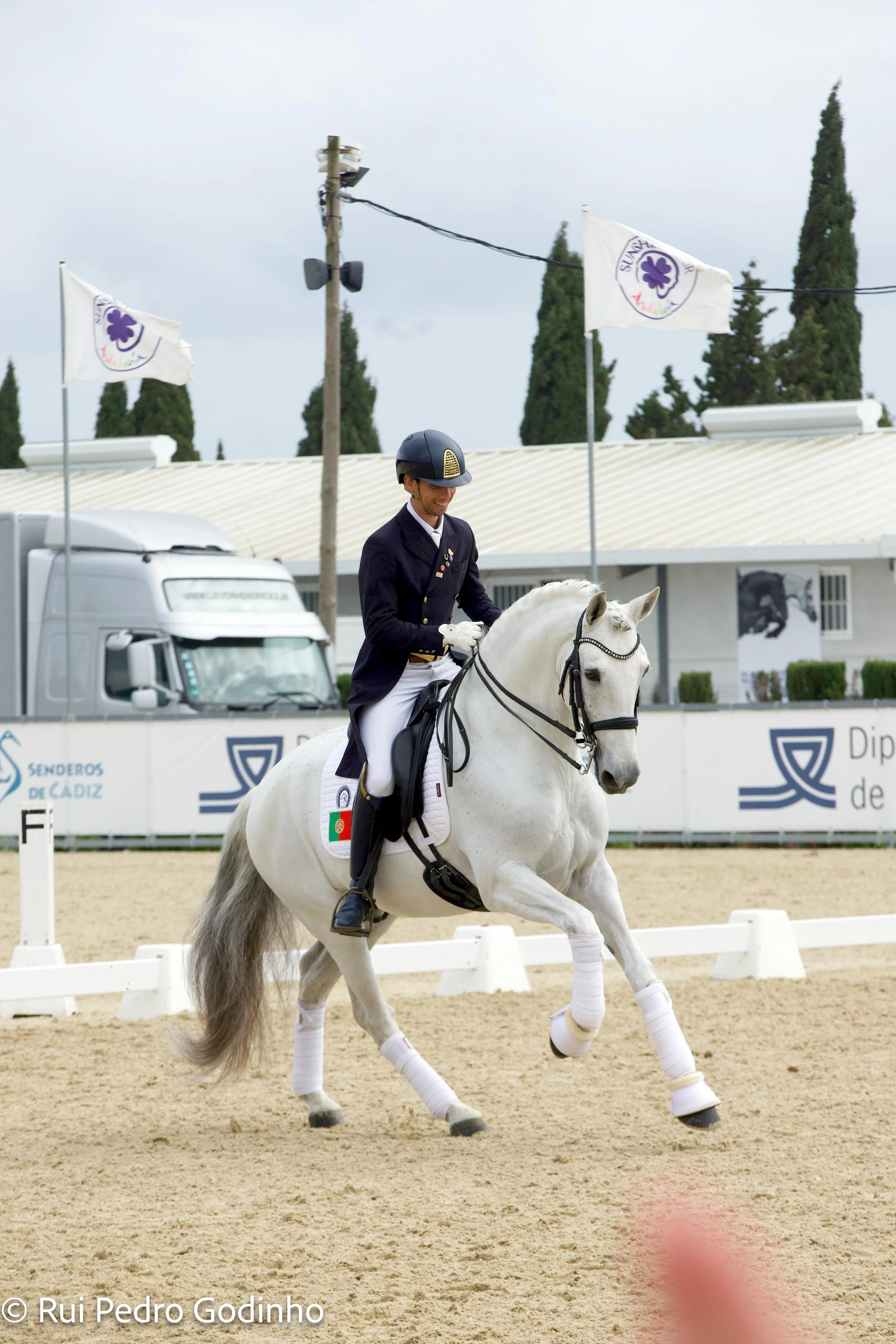 A male equestrian rider on a white horse during a dressage competition. The rider is wearing a dark jacket, white breeches, and a black helmet, and the horse is decorated with leg wraps and a black bridle.