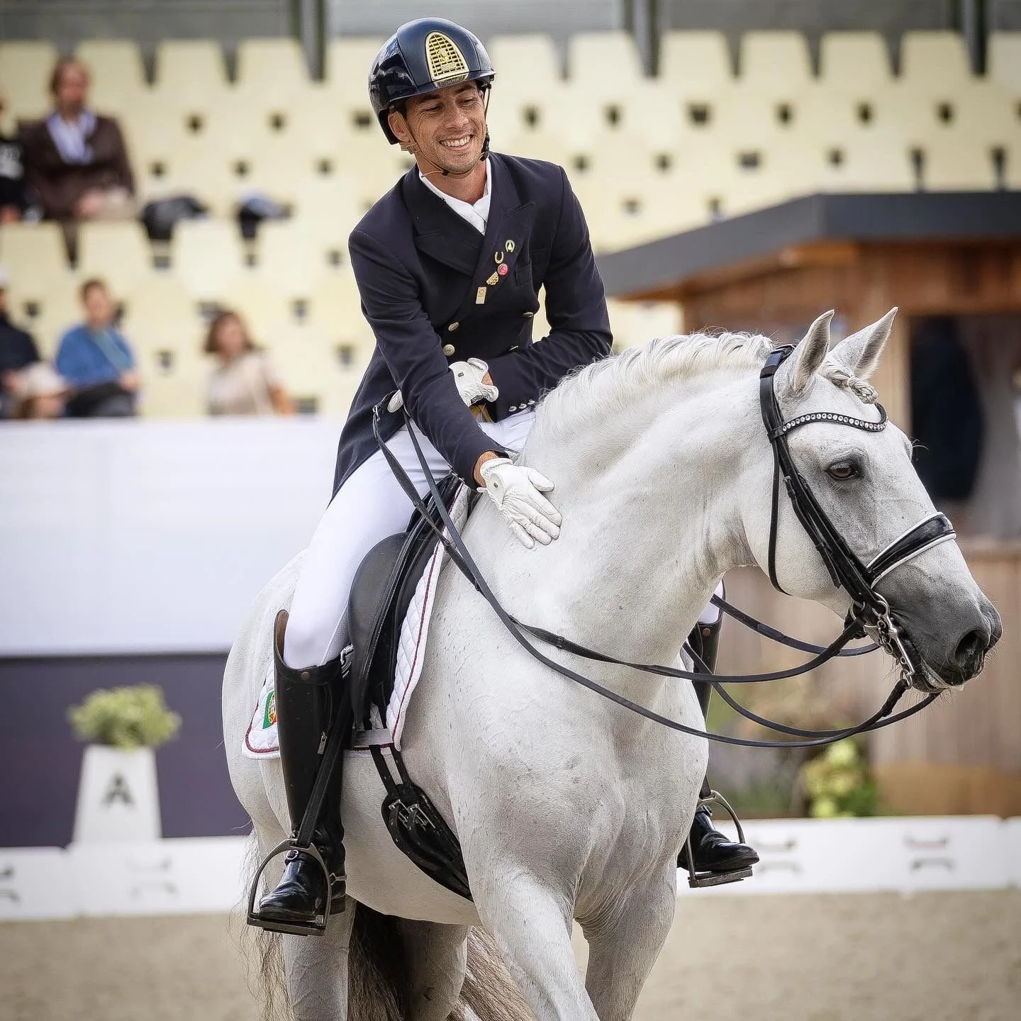 A smiling male equestrian in formal riding attire on a white horse during a competition.