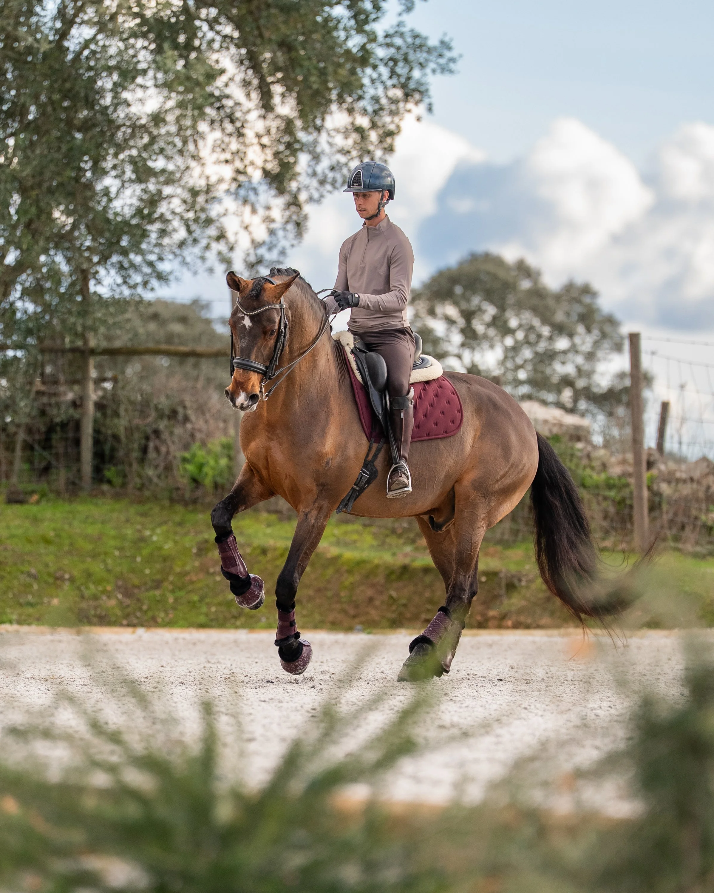 A person riding a Brown horse with black mane and tail in a riding arena. The rider is wearing a helmet, beige long-sleeve shirt, brown riding pants, and gloves. The horse has protective leg wraps and a maroon saddle pad. The sky is partly cloudy wit