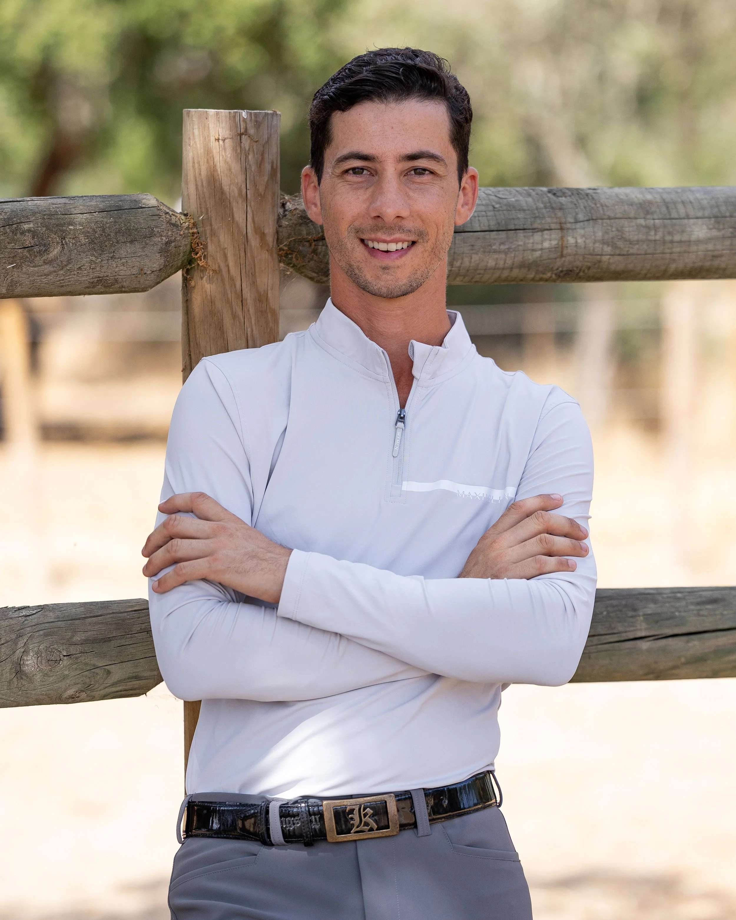A smiling man with dark hair and light skin, wearing a white zip-up sports shirt and gray pants, standing outdoors against a wooden fence, with trees in the background.