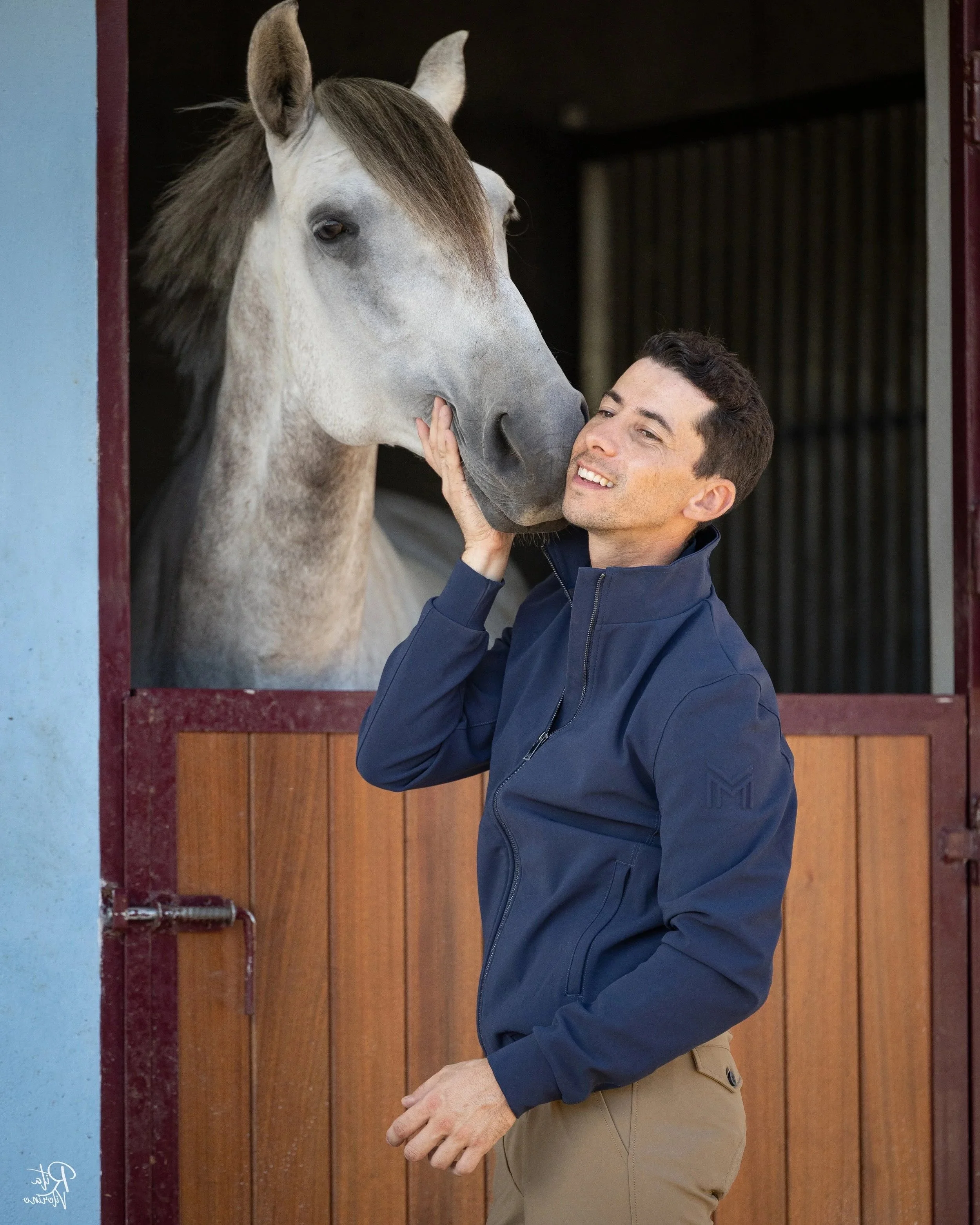 A man gently touching the nose of a gray horse through a stable window, both appearing happy and affectionate.