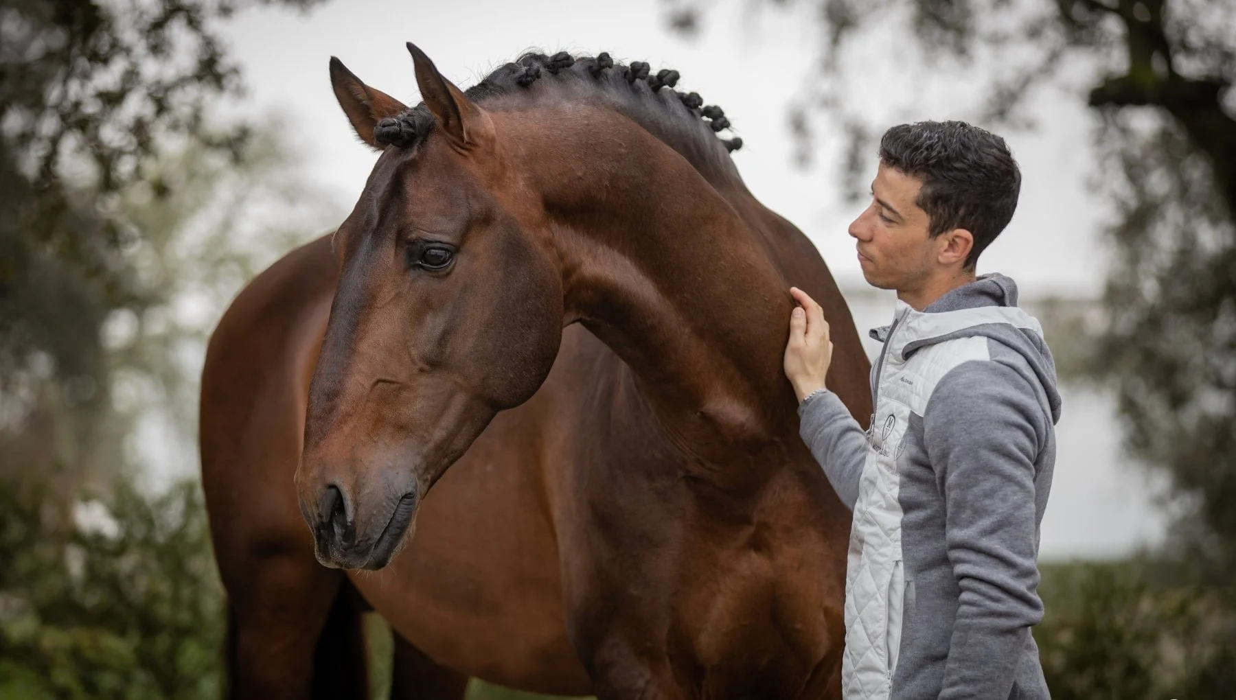 A man gently petting a brown horse outdoors with trees and cloudy sky in the background.