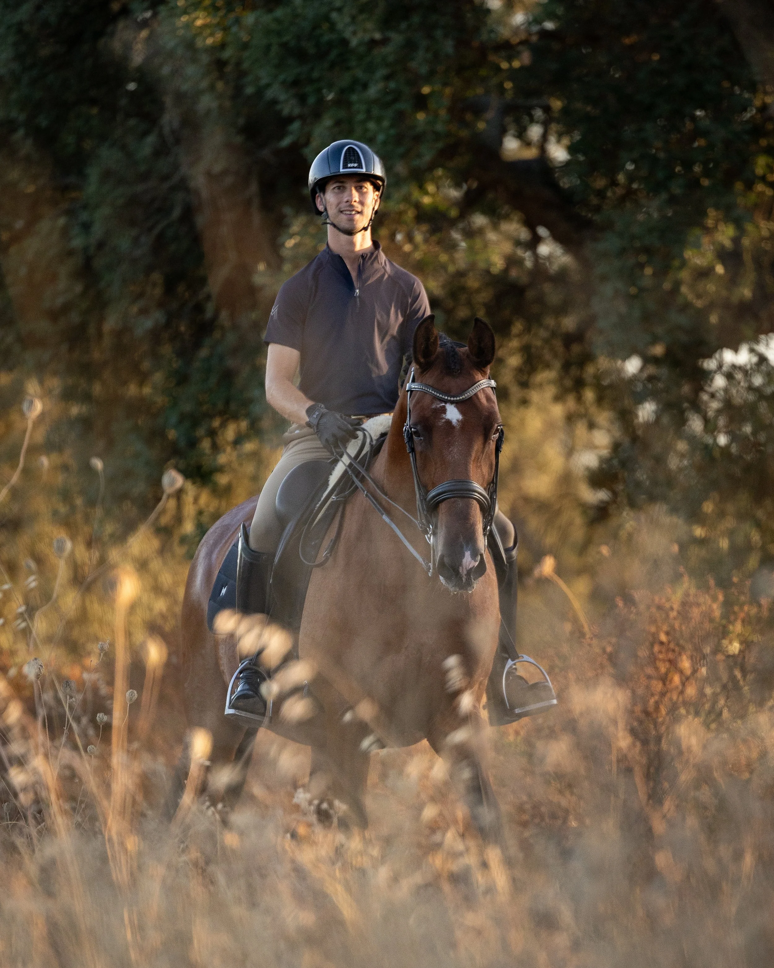 A man riding a brown horse through a nature trail at sunset. The man is wearing a black helmet, black shirt, and beige pants. The background features trees and golden light.