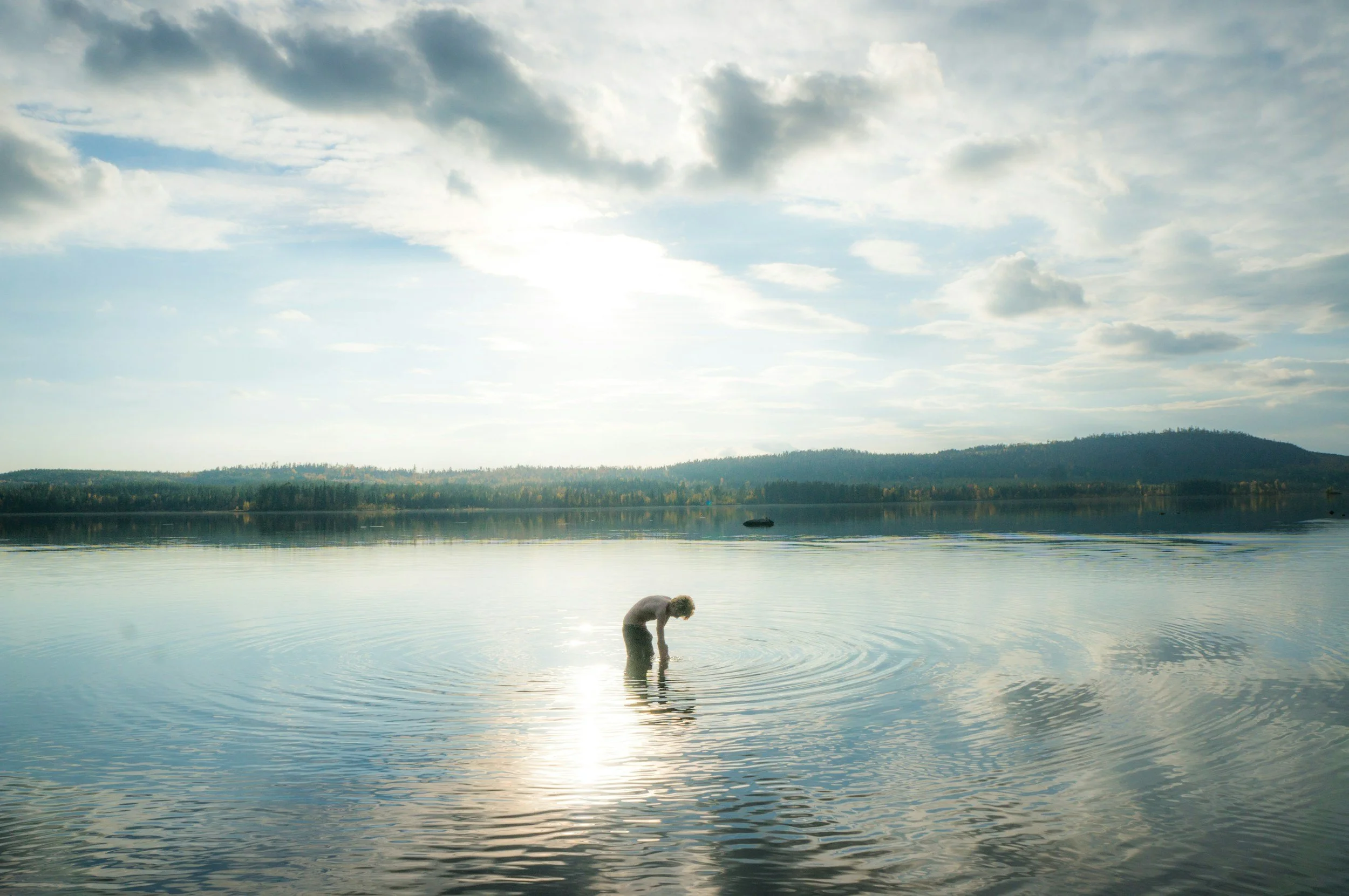 Person standing alone in shallow water, looking down as ripples spread outward across a calm lake under a wide sky.