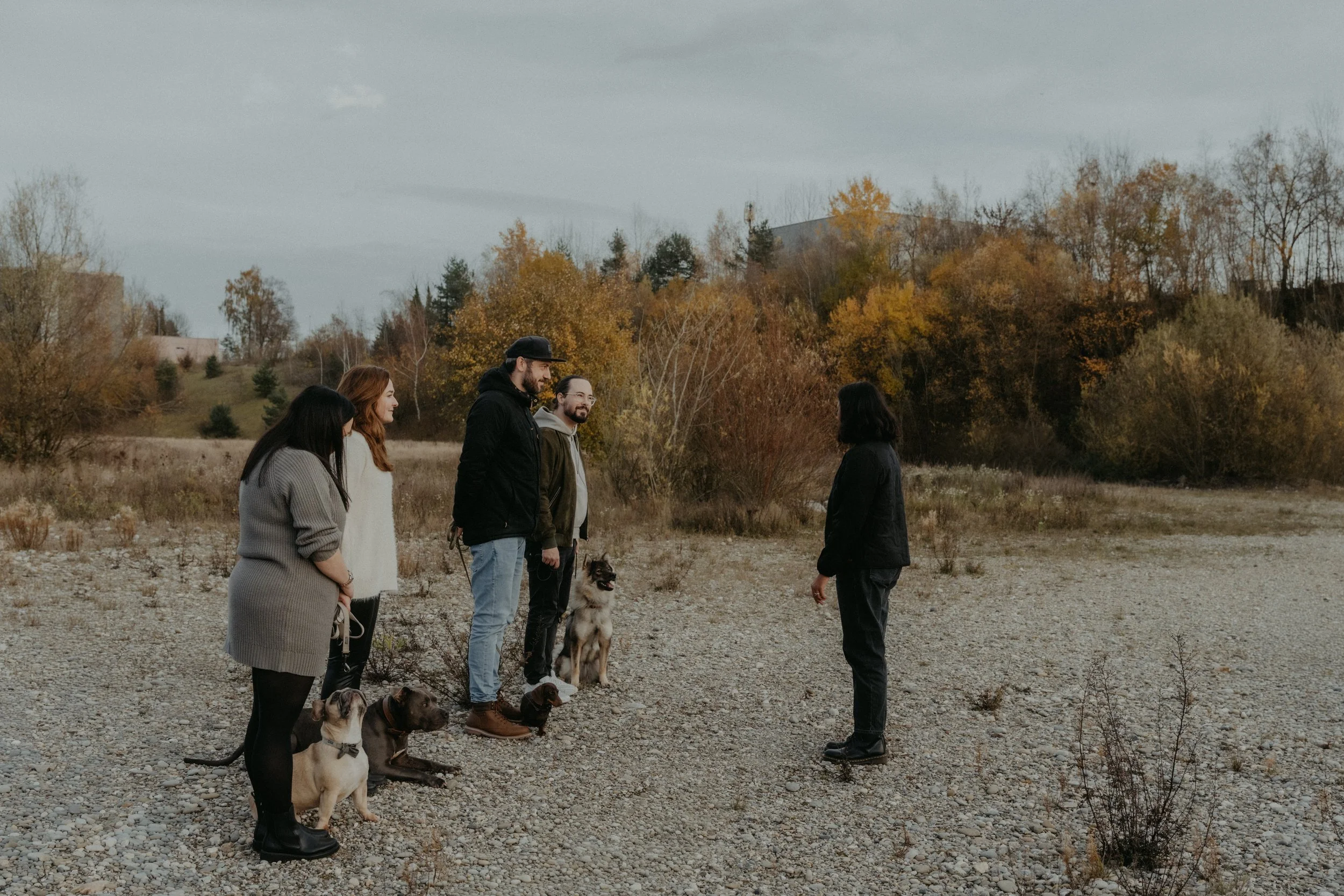 Fünf Menschen stehen in einer Reihe im Freien, mit drei Hunden, während sie einem anderen Menschen zuhören, im Herbst, auf einem kiesbedeckten Feld mit Bäumen und Hügel im Hintergrund.