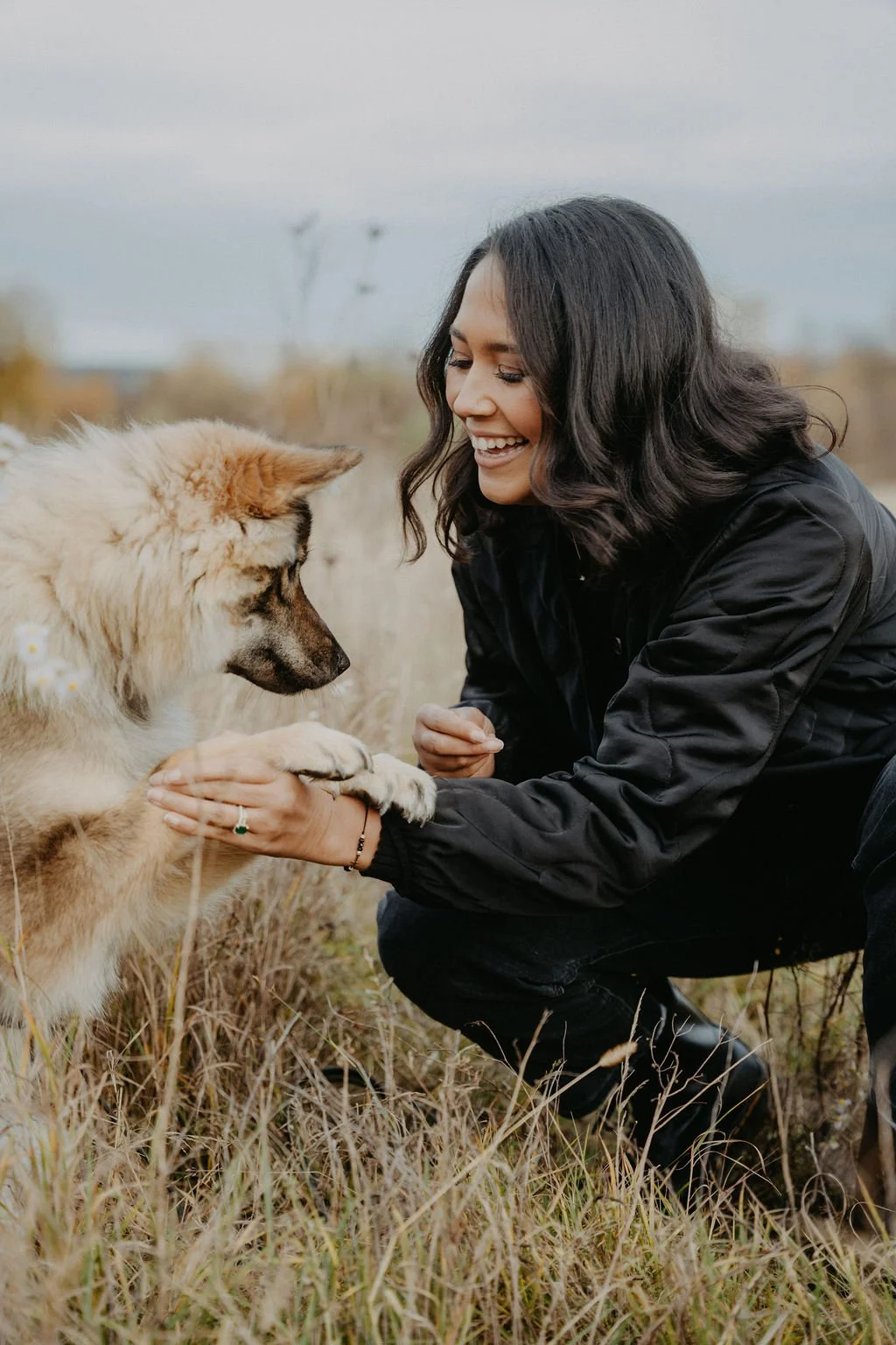 Eine lachende Frau im schwarzen Jacke hält die Pfote eines Hundes in einem Feld, unter einem grauen Himmel.