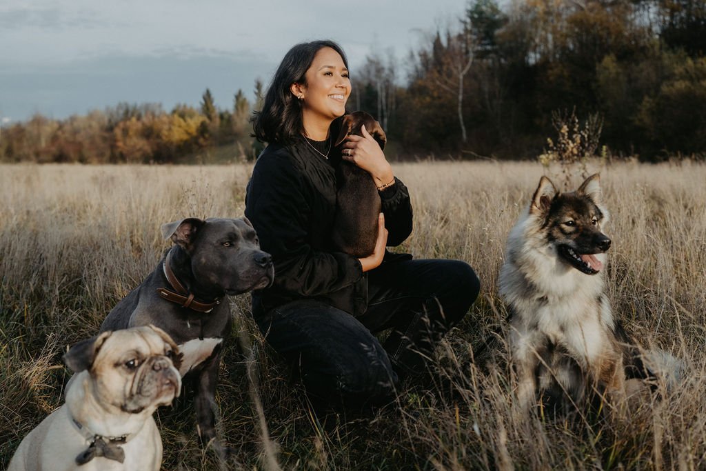 Eine Frau sitzt im Feld umgeben von vier Hunden und hält zwei davon im Arm, während sie lächelnd in die Ferne schaut.