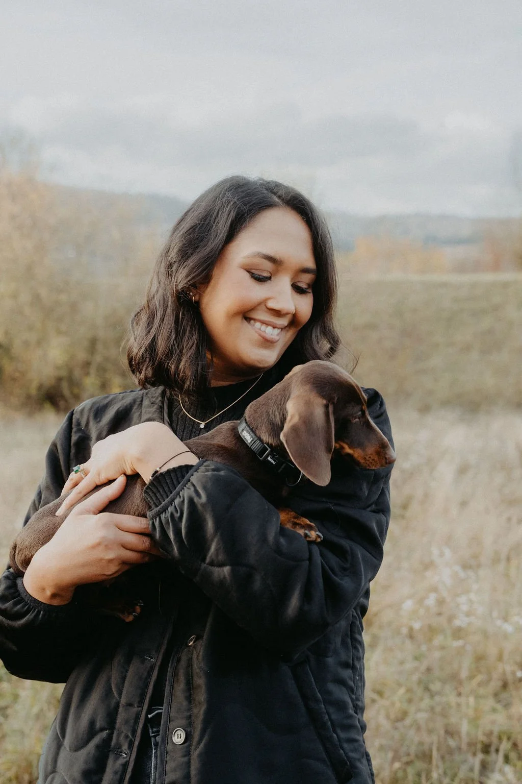 Junge Frau hält einen braunen Hund in den Armen, lächelt in die Richtung des Hundes, draußen im Feld mit Bäumen und Bergen im Hintergrund bei bewölktem Himmel.