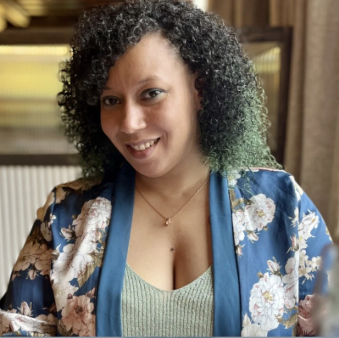 A woman with curly, black and green hair smiling and wearing a floral blue jacket over a light green top, indoors with wood and yellow patterned background.