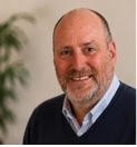 A portrait of a middle-aged man with a bald head, beard, and smiling, wearing a dark sweater over a collared shirt, standing in front of a green plant.