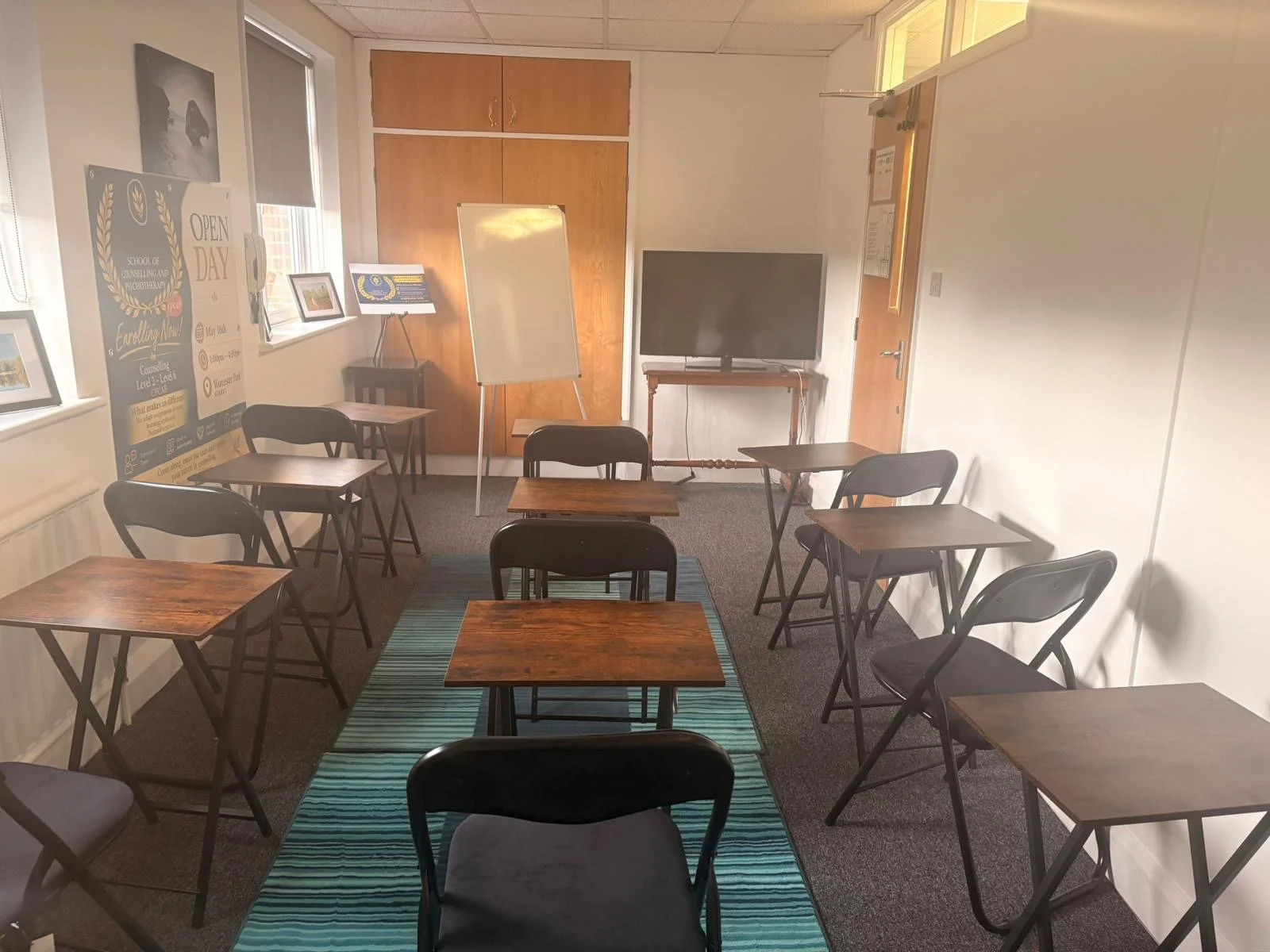 A small conference or classroom room with several wooden tables and black chairs arranged around a blue striped carpet. There is a television on a stand, a whiteboard on an easel, and windows with blinds on the left wall.
