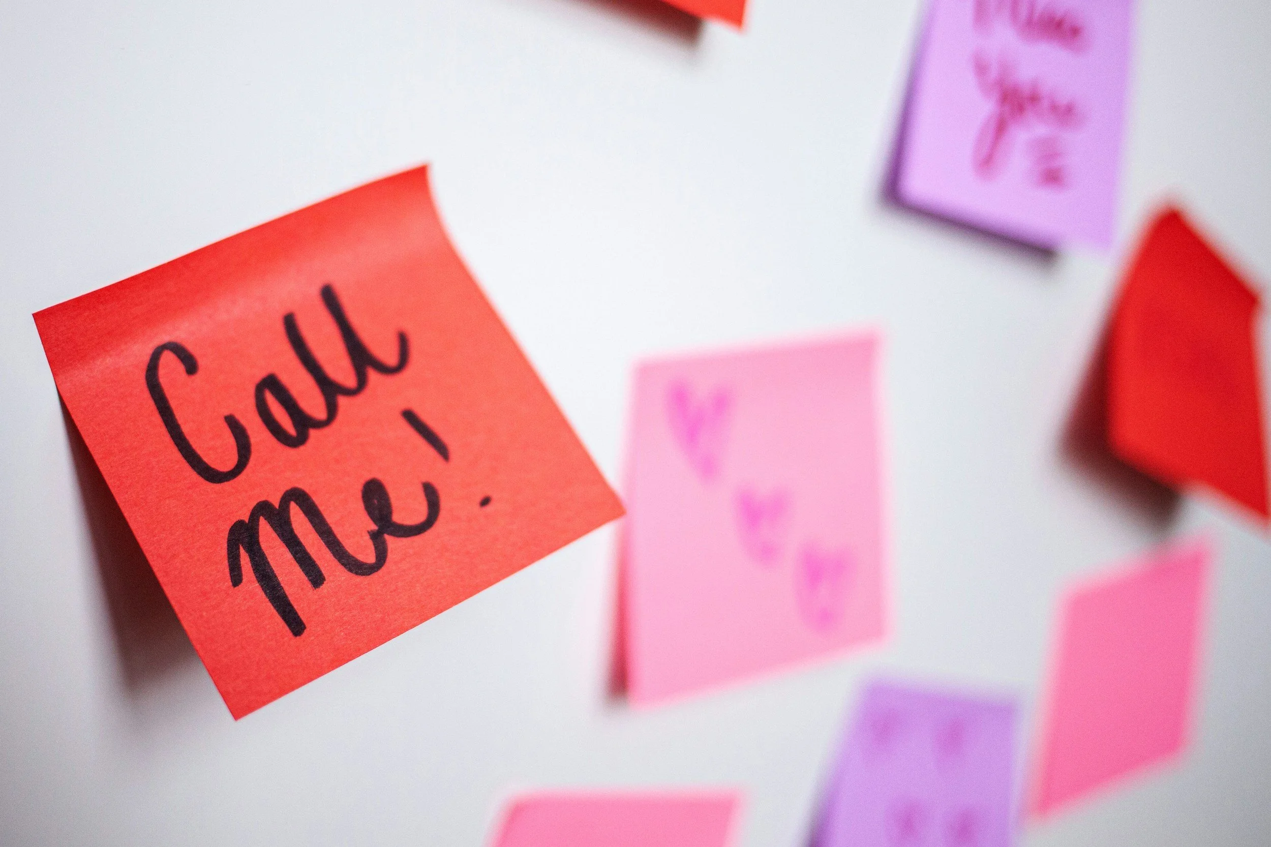 A close-up of a red sticky note with the handwritten message "Call me!" on a whiteboard, surrounded by other pink and purple sticky notes.
