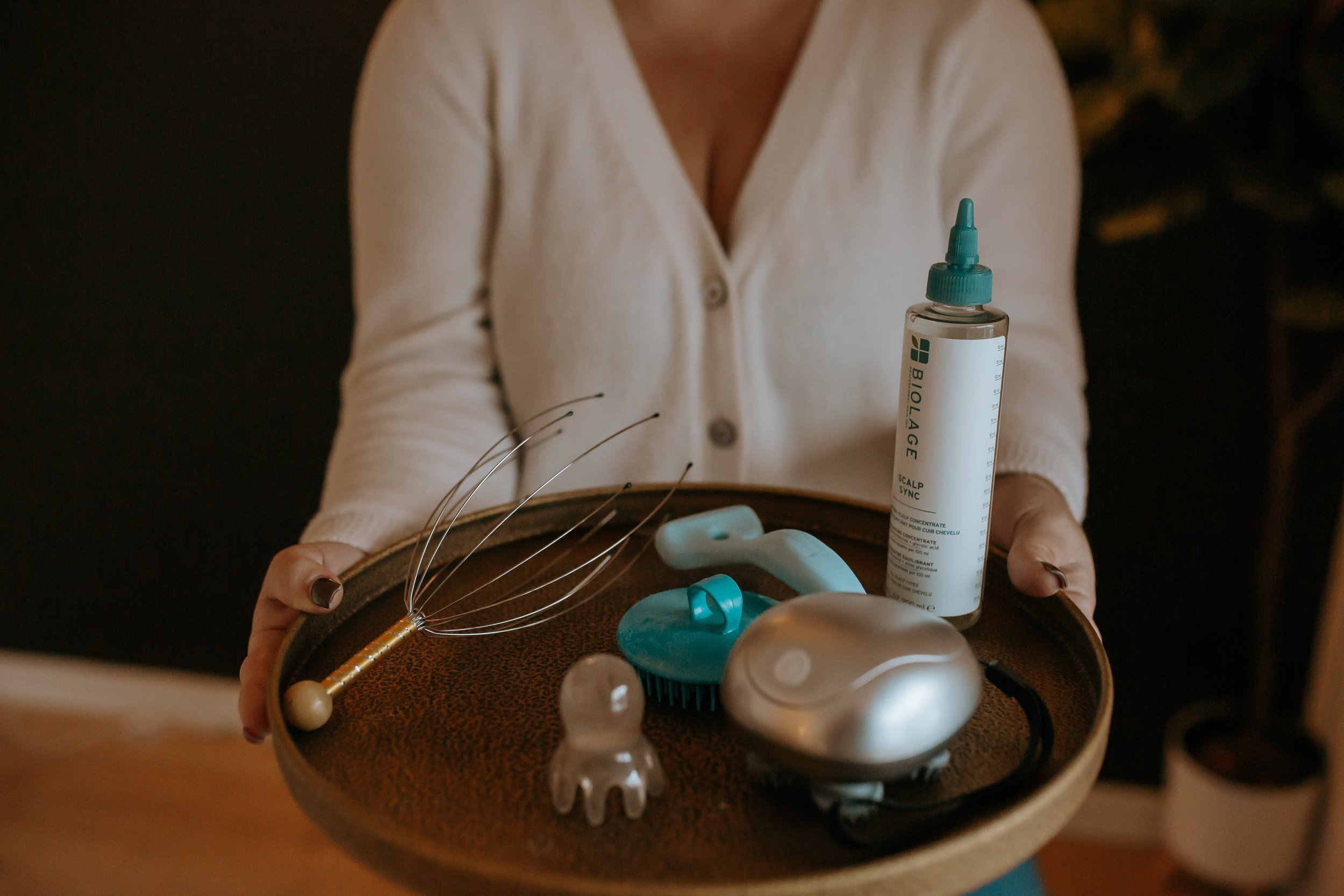 Person holding a round tray with scalp massage tools, a bottle of scalp serum, and a facial massage device.