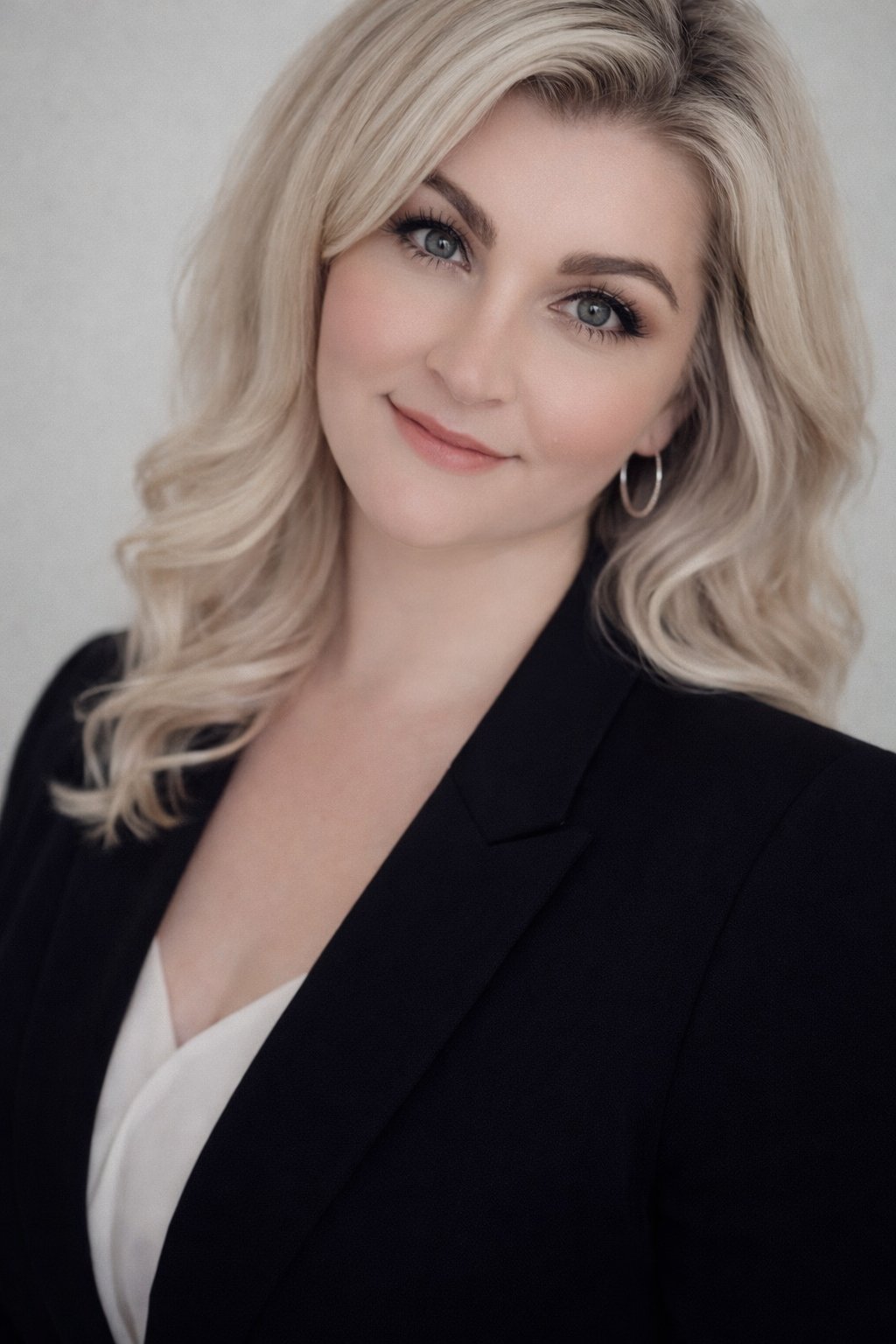Portrait of a woman with blonde, wavy hair wearing a black blazer and earrings, smiling softly against a plain background.